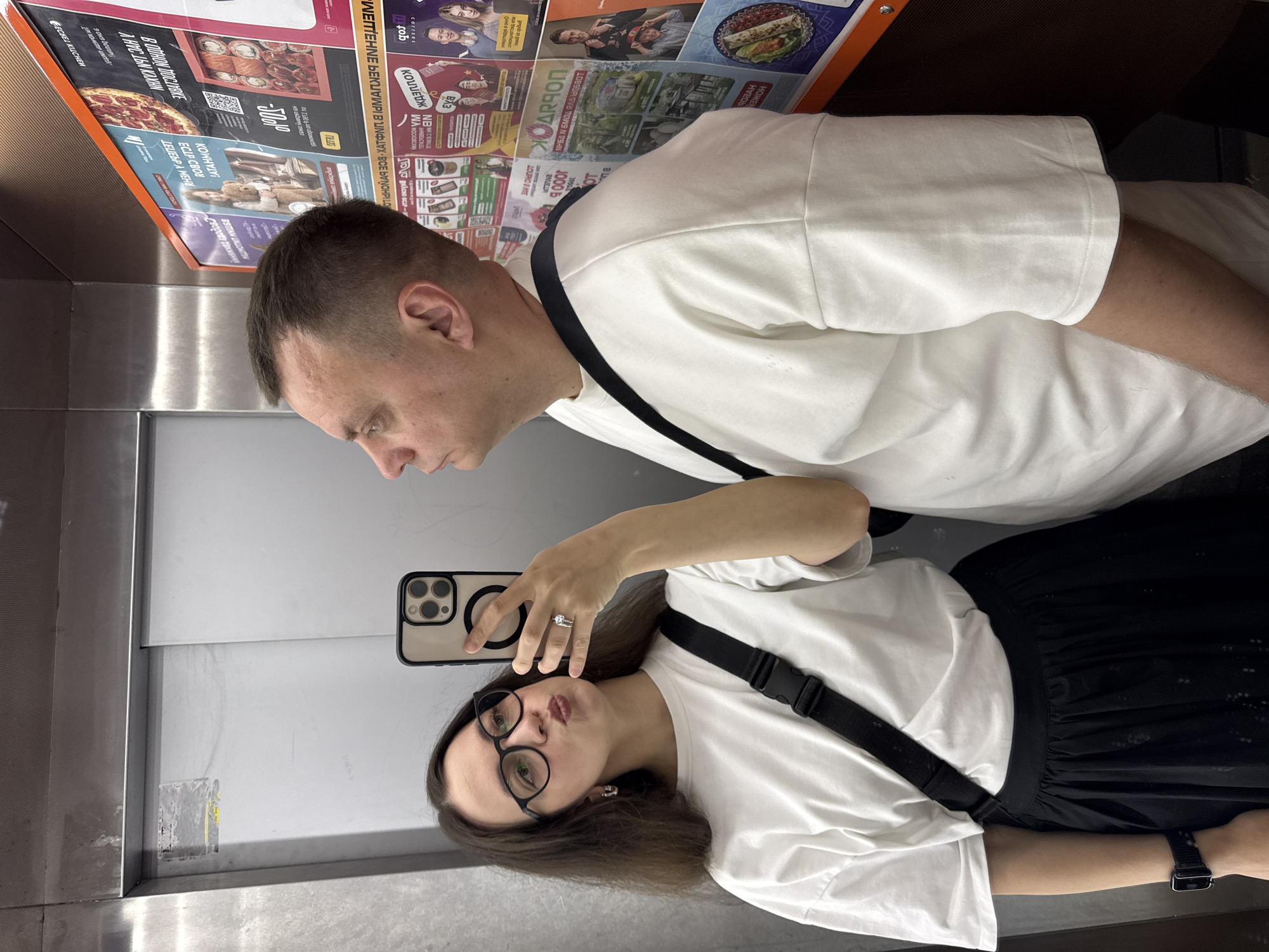 This image shows a selfie of a woman and a man inside an elevator. The woman is in the foreground, holding a smartphone to take the picture. She is wearing glasses, a white t-shirt, a black skirt, and a black strap bag. The man is behind her, also wearing a white t-shirt and a black strap bag. In the background, there is a bulletin board with advertisements in Russian. Based on the advertisements, the location is Nizhny Novgorod, Russia. The walls of the elevator are silver.