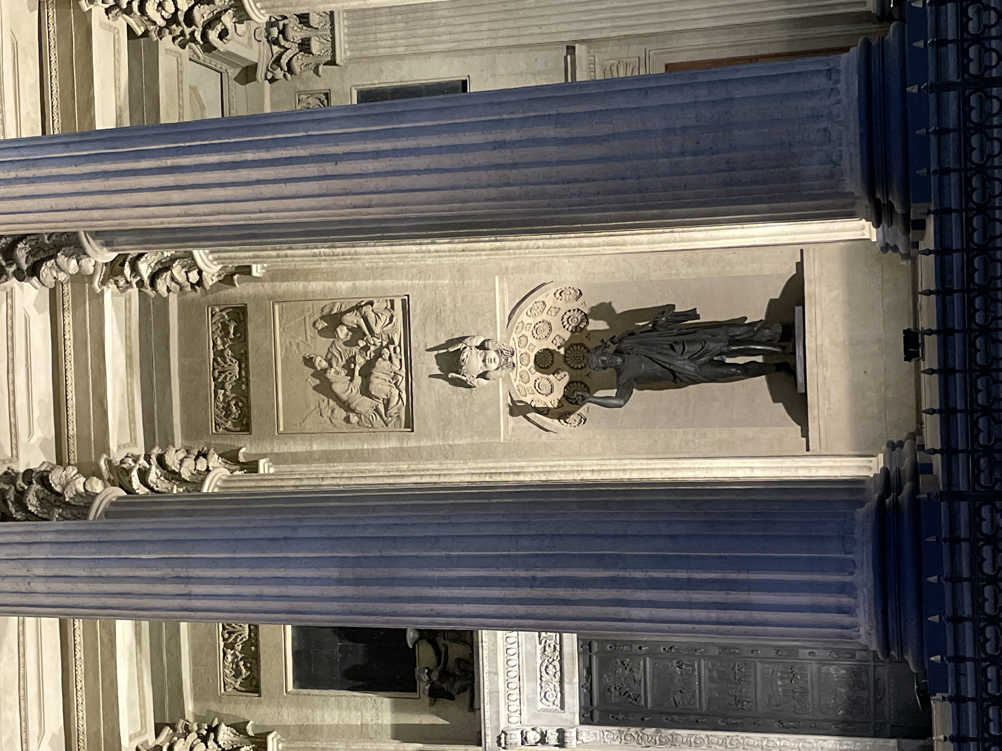 This detailed image captures a section of the Kazan Cathedral facade in Saint Petersburg, Russia, at night, illuminated by artificial lighting. The scene is dominated by massive, fluted Corinthian columns of light grey stone, exhibiting a blue tint from the cool illumination. Behind the columns, a wall features decorative elements, including a prominent arched niche housing a dark bronze statue of a male figure. The statue, likely depicting Saint Andrew the First-Called, is draped in robes and appears to hold a staff or cross in his left hand, standing on a pedestal within the intricately decorated niche that features patterned rosettes in its semi-circular upper section. Above this niche, a rectangular bas-relief panel depicts a group of multiple figures engaged in a scene. To the viewer's left, partially obscured by a column, a section of a large, ornate bronze door with sculpted relief panels is visible, above which a small, dark rectangular window is present. On a decorative ledge beneath this window, a small, kneeling cherub-like sculpture is positioned, holding an object. The artificial lighting, primarily from below, casts dramatic shadows, accentuating the architectural details, the fluting of the columns, and the three-dimensional quality of the sculptures and reliefs. A black, decorative metal fence with pointed finials runs across the bottom of the frame, partially visible in the foreground.