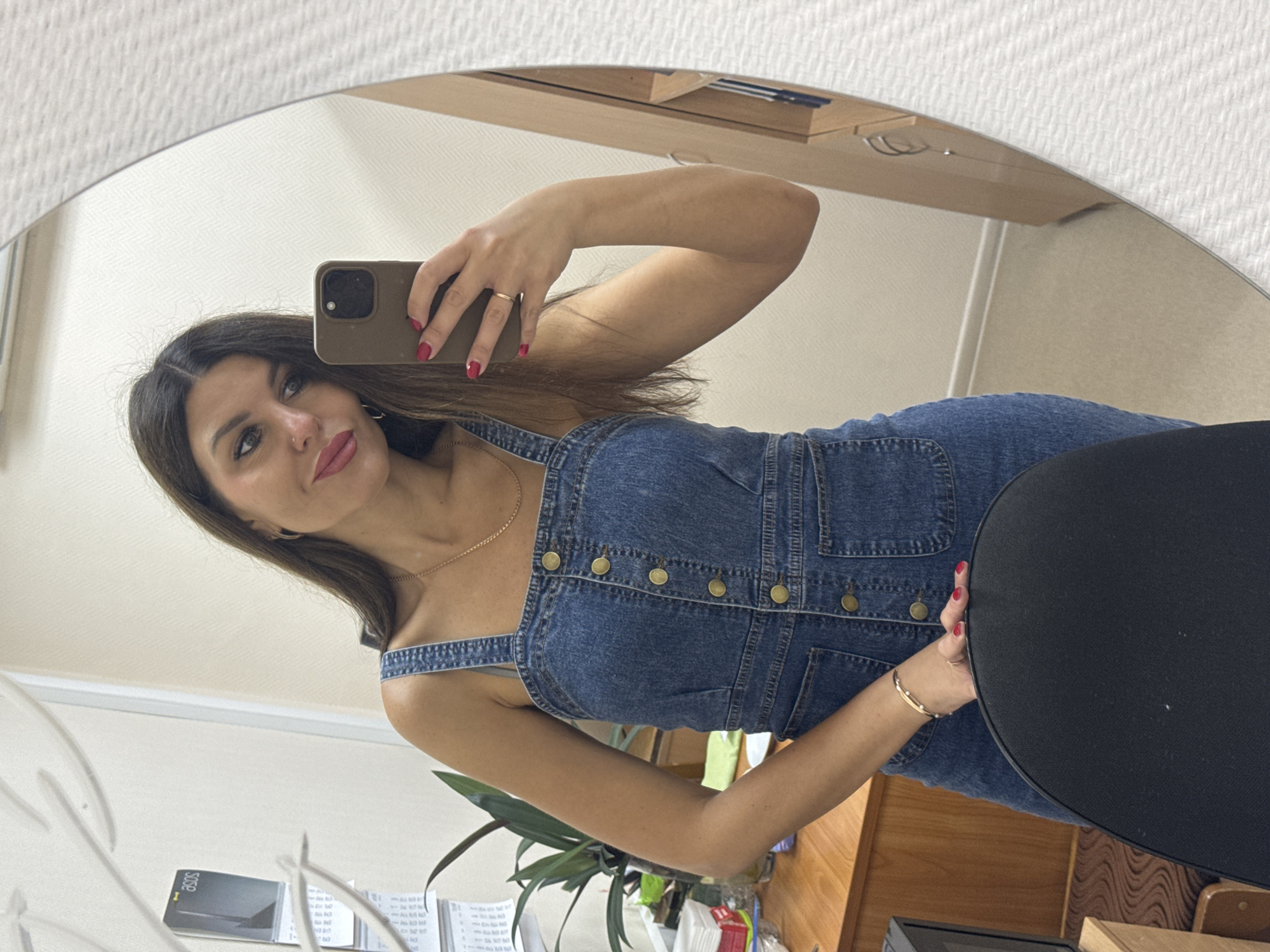The image shows a woman in a denim dress taking a selfie in a mirror. She has long brown hair and is wearing makeup. She is holding a smartphone in her right hand and her left hand is resting on a black chair. In the background, there is a desk with a plant and some papers. The scene appears to be indoors, possibly an office or a home. The metadata indicates the image was taken in Chaikovsky, Russia.
