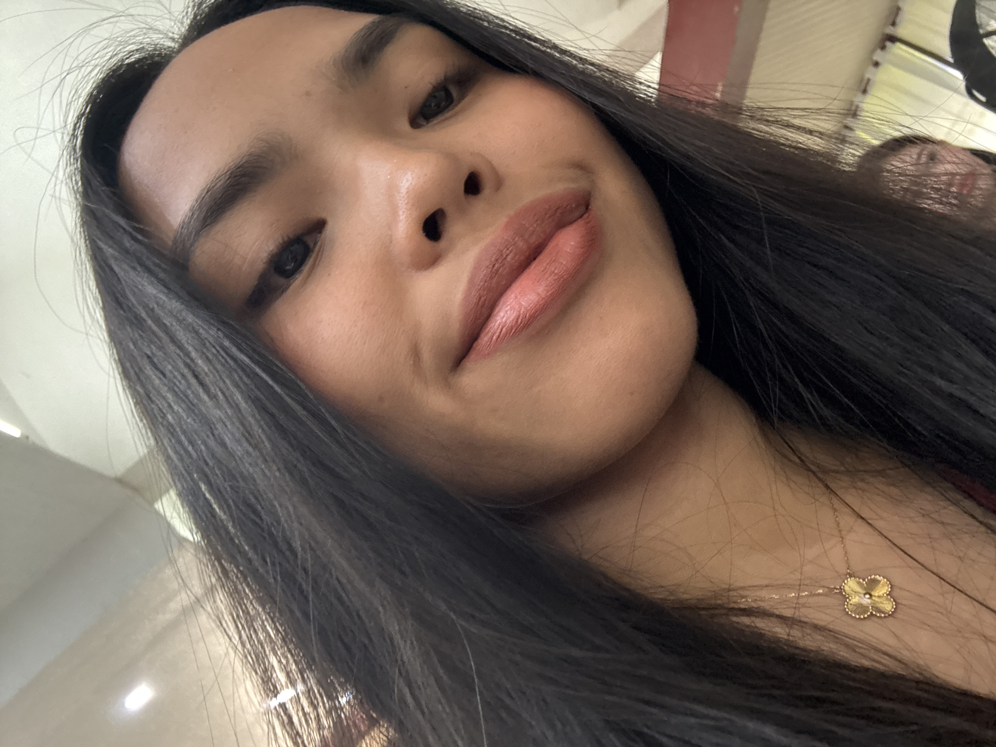 A medium shot selfie of a young woman is captured from a low angle. She has long, dark hair that is styled to partially obscure her face. Her skin tone is medium to tan, and she is wearing makeup, including lipstick in a peachy-pink shade. A delicate gold necklace with a four-leaf clover pendant is visible on her neck. The background is blurred, but appears to be an indoor setting with bright overhead lighting and some muted colors, possibly indicating an office or retail environment. The image does not provide any specific visual cues that would allow for the identification of its location within Davao City, Philippines.