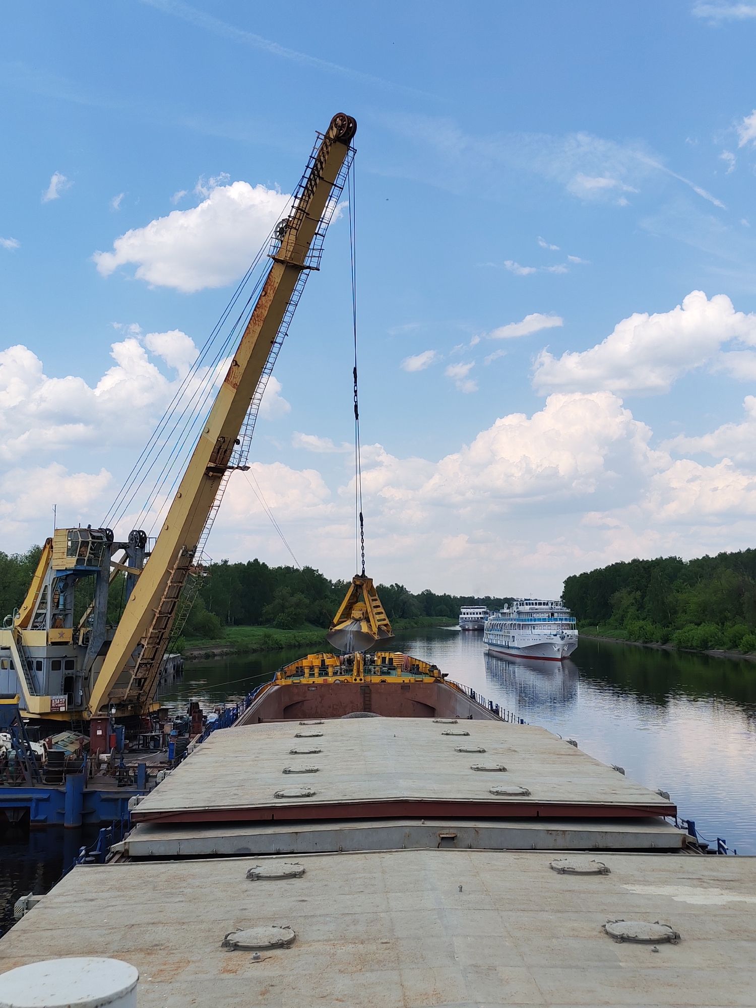 The image shows a river scene in Yakhroma, Russia. In the foreground is the deck of a barge, with a large yellow crane positioned on its side. The crane has a clamshell bucket suspended over the barge. In the background, two passenger cruise ships are visible on the waterway. The river is flanked by green trees on both banks. The sky is blue with scattered white clouds.