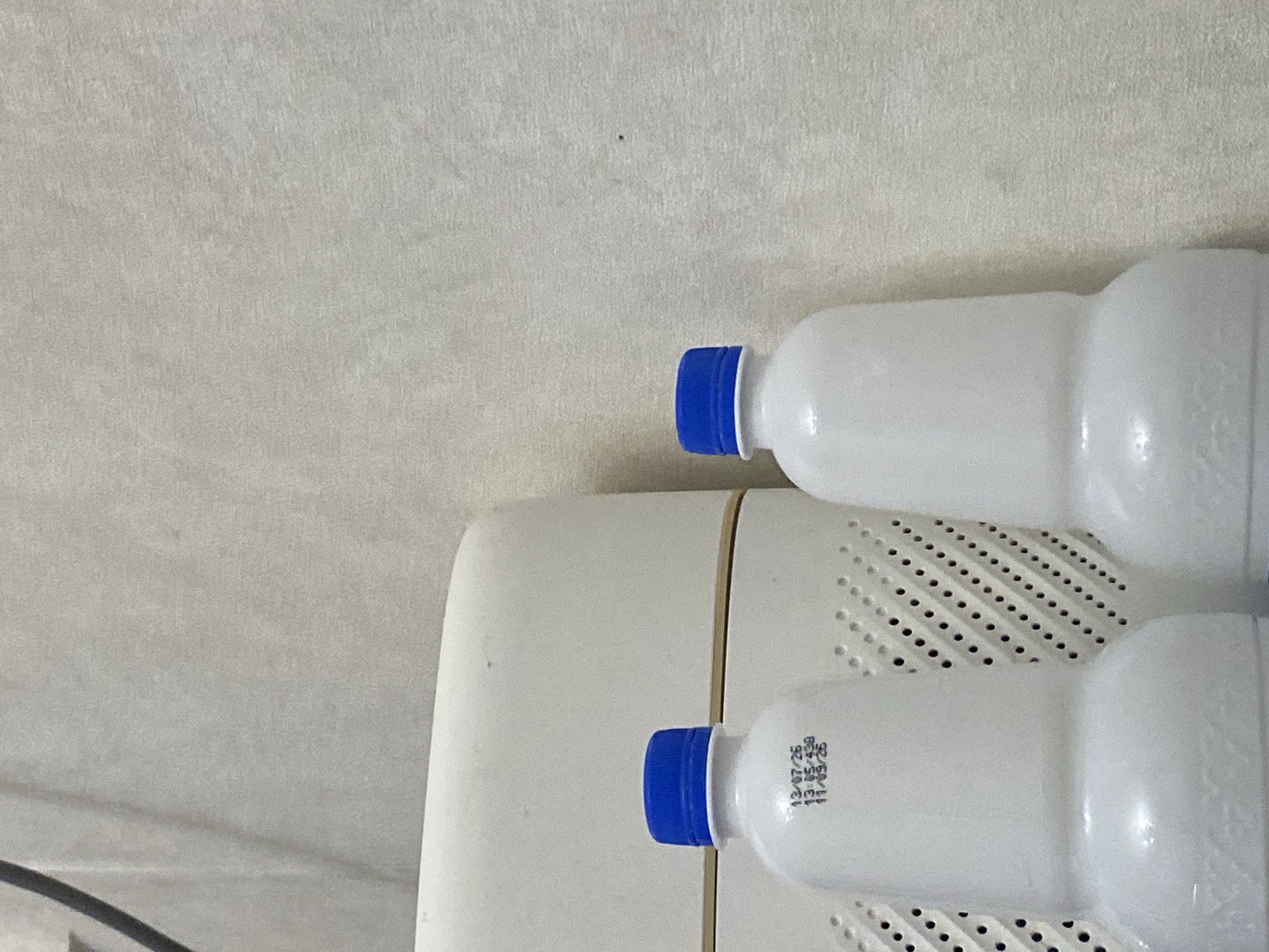 Two white plastic bottles with blue caps are positioned in front of a white, rectangular object with a perforated surface. The bottles have black lettering printed on them. The bottle on the left displays the following text: "13/07/25", "13:05/439", and "11/05/25". The background is a textured, light-colored wall. The scene is indoors. No specific location in Bishkek City, Kyrgyzstan is identifiable.