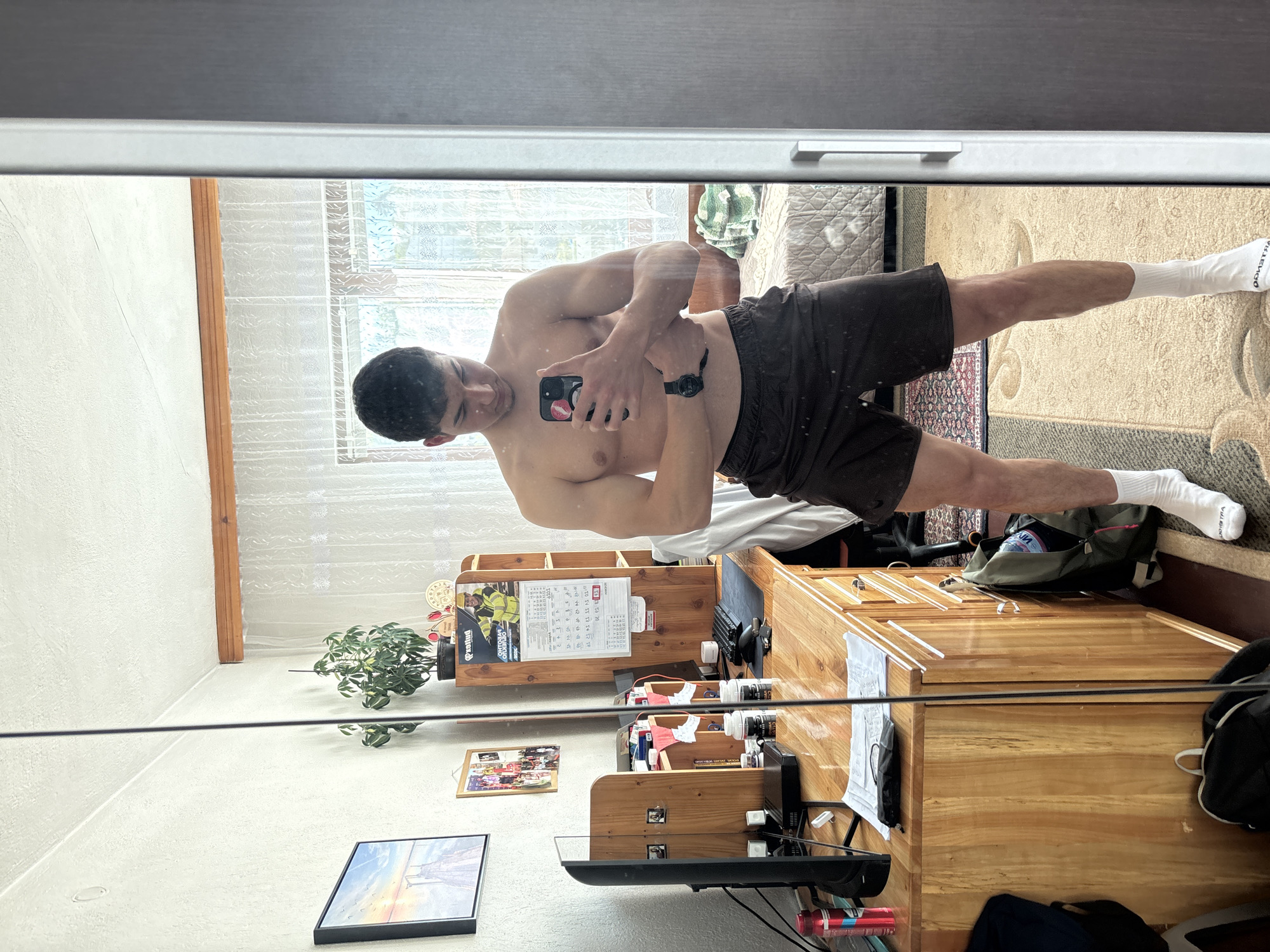 The image captures a mirror selfie taken by a young man indoors. He is shirtless, showcasing visible muscle definition, and wears dark shorts and white socks. He holds a black smartphone with a red kiss-mark sticker on its case, actively taking the picture, and a black smartwatch is visible on his left wrist.
The reflection in the mirror reveals a domestic setting, likely a bedroom or living area. The room features light-colored walls, a window covered with sheer white curtains, and a patterned beige rug covering a significant portion of the floor. Various wooden furniture pieces are present, including a large dresser or cabinet with multiple drawers and a wooden shelf unit. On the shelf unit, a calendar with Cyrillic script, displaying "Октябрь" (October), is visible, along with a small potted plant and various bottles or jars. A monitor or television screen, a framed picture, and a green and black backpack are also seen on or around the furniture. A bed with patterned, quilted bedding is partially visible in the background.
The lighting suggests it is daytime, with natural light entering the room. Visible text includes "VILLBHO" on one of the white socks and a bag, "LENGO" on a label on the shelf, and "Октябрь" on the calendar. The scene depicts a casual moment in what appears to be a personal space, consistent with a home environment in Zlatograd, Bulgaria.