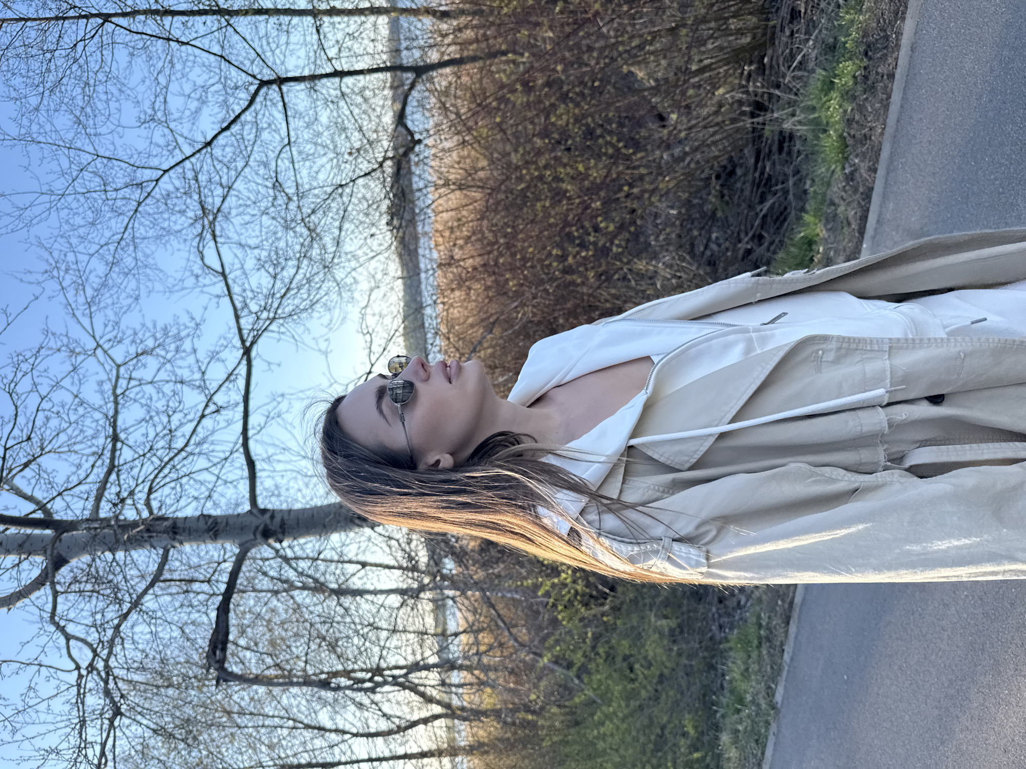 A young woman with long brown hair and sunglasses is standing outdoors on a paved path. She wears a beige trench coat over a white shirt or jacket. Her head is tilted back, and she appears to be looking up. In the background are bare trees and a body of water. To the right is a dense bush. The location appears to be a park or recreational area near a body of water, but the context of Białobrzegi, Poland is not clear from visual information.