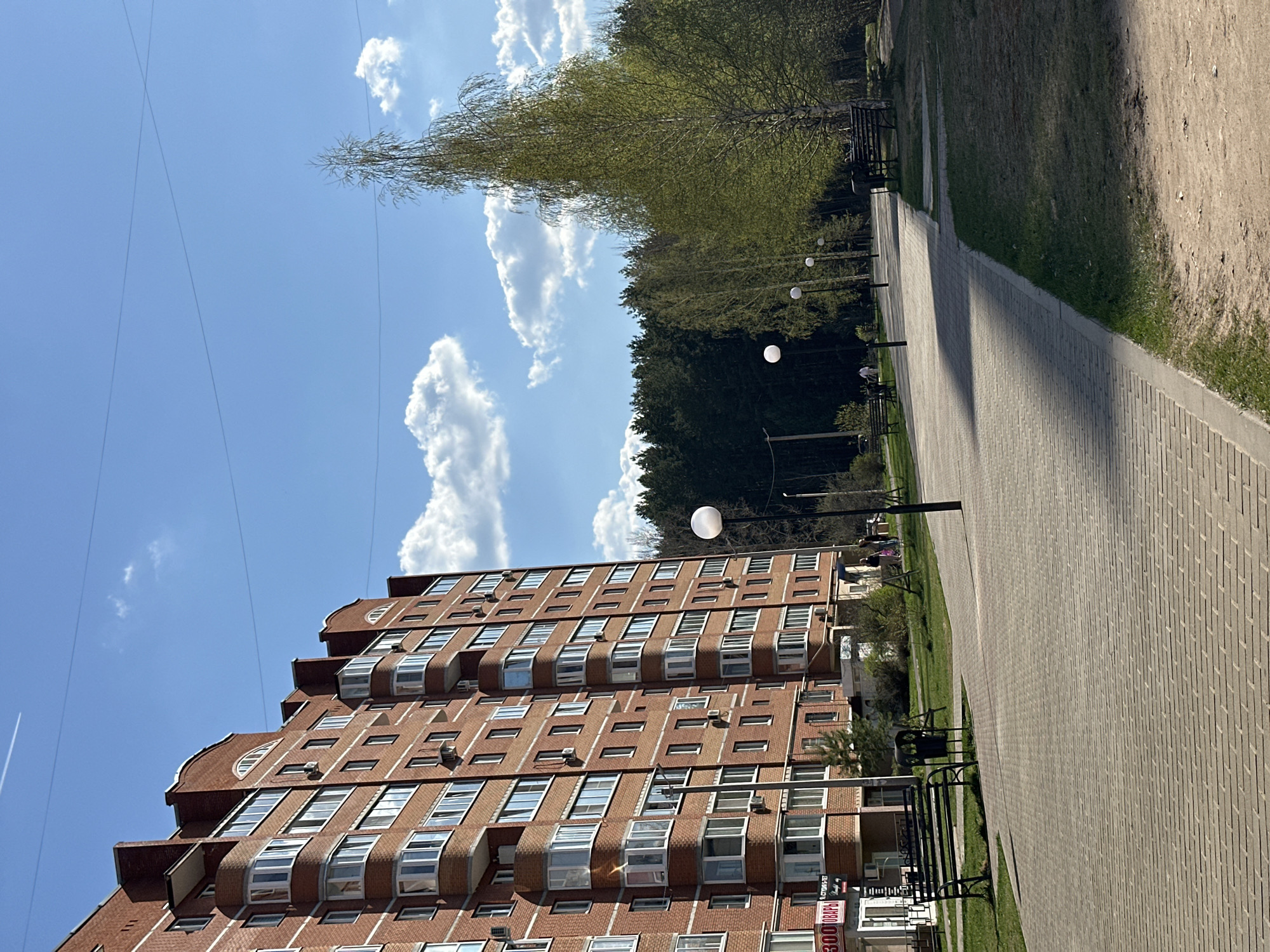 The image depicts an urban scene in Chaikovsky, Russia, featuring a multi-story brick apartment building with distinctive stepped facades and numerous windows, suggesting a residential area. The building is situated next to a paved walkway lined with trees and benches, forming a public space or park. Spherical streetlights are spaced along the walkway. The sky is blue with scattered white clouds. A faint contrail from a high-altitude aircraft is visible in the sky. No people are clearly discernible in the frame.