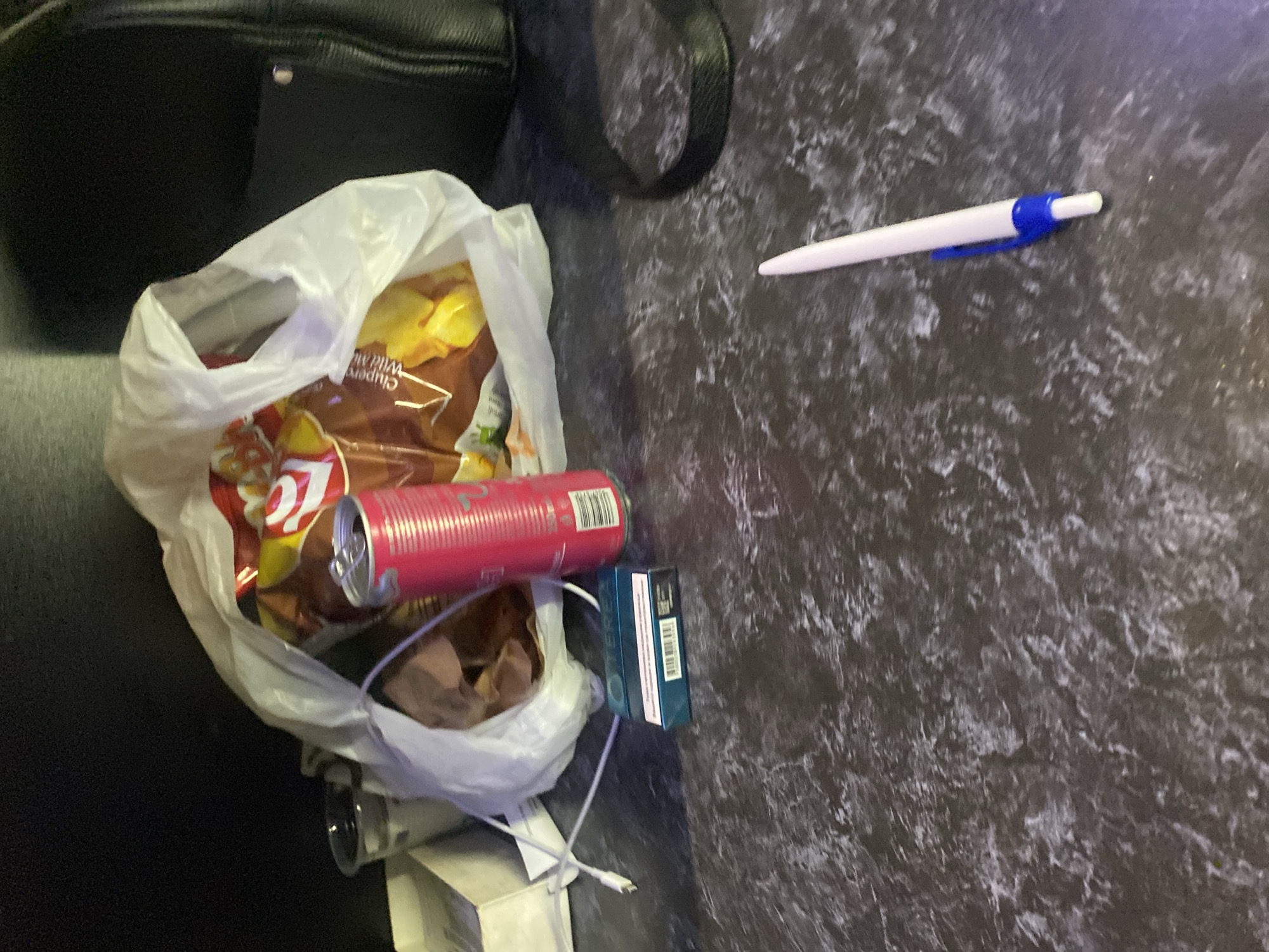 The image depicts a dark, cluttered surface, possibly a table, with various objects scattered across it. In the background, a black bag is partially visible. In the mid-ground, a white plastic bag is filled with items, including what appears to be a bag of snacks with red and yellow packaging. A pink soda can is positioned in front of the plastic bag, and next to it is a small blue box with white lettering, possibly cigarette packaging. A white charging cable is also visible, running from the left side of the image towards the plastic bag. In the foreground, a white pen with a blue clip rests on the surface. The surface itself has a mottled, dark gray and white pattern. The scene is dimly lit, creating shadows and obscuring some details. The overall impression is one of casual disarray. There is no discernible location in the city of Beloslav, Bulgaria, or any indication of people or actions.