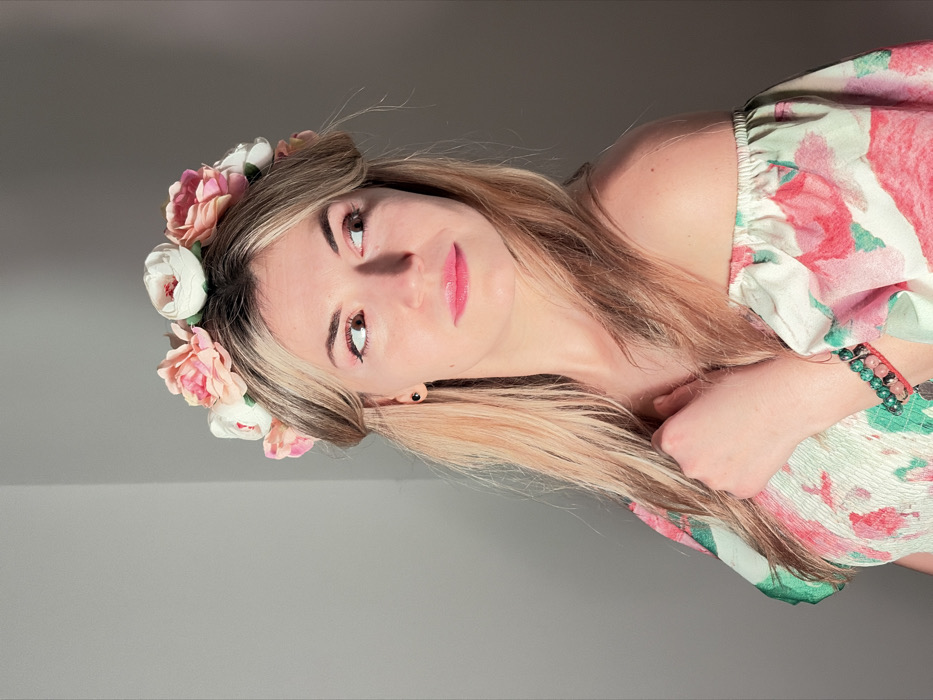 The image depicts a young adult female, fair-skinned with long blonde hair, standing against a plain light grey wall. She is wearing a flower crown composed of pink and white artificial peonies and an off-the-shoulder garment with a floral pattern in pink, green, and white. A black stud earring is visible in her left ear. Her lips are coated with pink gloss. On her left wrist, she wears two bracelets: one with green beads and another with reddish string. Her right hand is positioned near her upper chest/collarbone area. Her gaze is directed towards her right (viewer's left). The lighting illuminates her from the left, casting subtle shadows. This scene is located in the city of Budva, Montenegro, within a non-descript indoor setting.