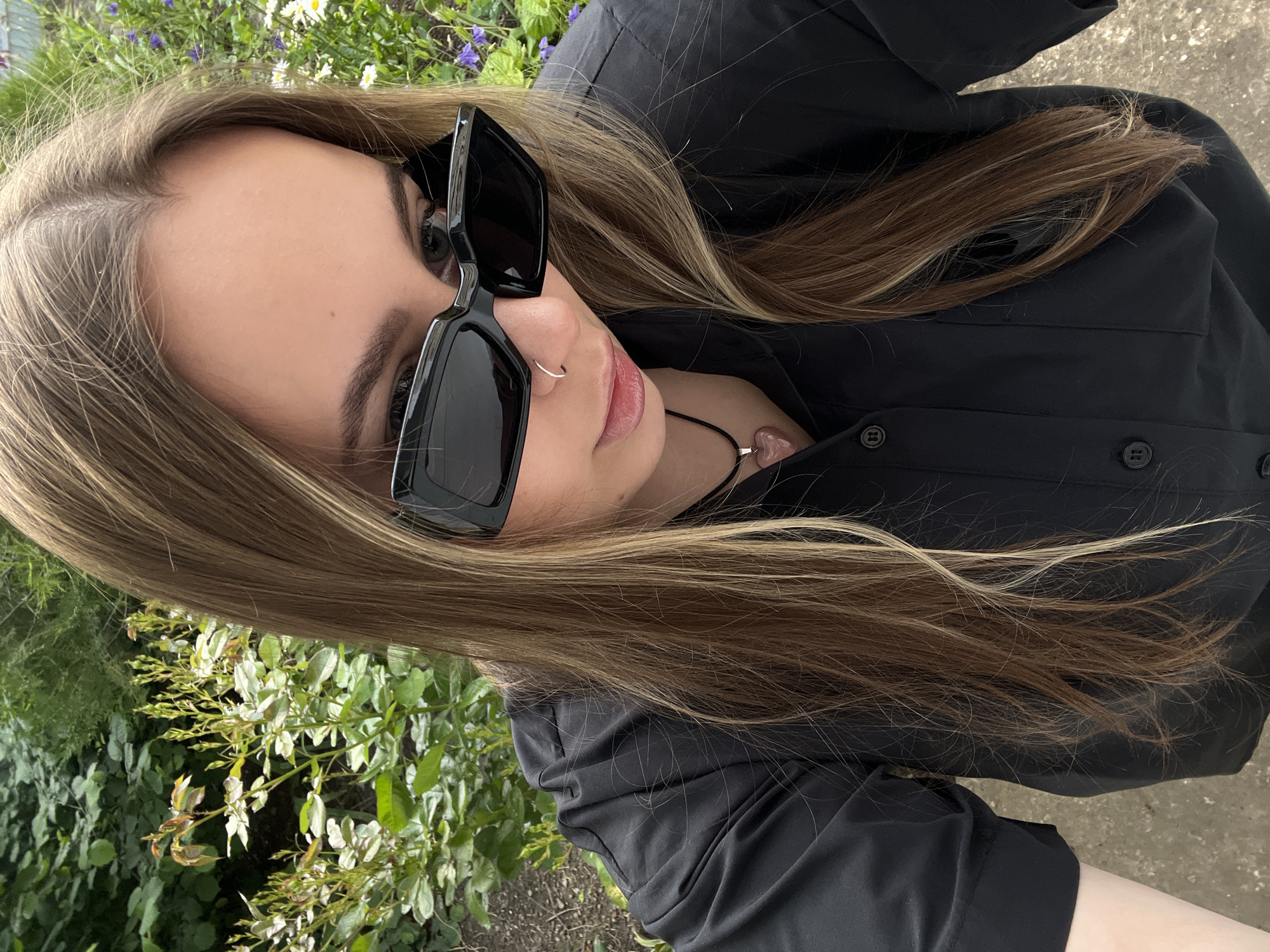 A young woman is captured in a close-up, angled shot, appearing to be taking a selfie or being photographed from a low perspective. She has long, straight hair, predominantly light brown with distinct blonde highlights, framing her face and resting over her shoulders. She wears large, square-framed black sunglasses that cover her eyes and much of her upper face. A small silver hoop nose ring is visible on her right nostril. Around her neck, she has a black cord necklace adorned with a light pink, heart-shaped pendant. She is dressed in a black collared shirt or jacket, which features visible buttons down the front and pocket details.
The background is an outdoor setting filled with green foliage, including various leaves and small branches. Scattered among the greenery are small white daisy-like flowers and delicate purple blossoms, suggesting a garden or park environment. The lighting is soft and even, indicating it is daytime, possibly overcast or the subject is in a shaded area. No specific activity beyond posing for a picture is discernible, and her expression is mostly obscured by the sunglasses, conveying a casual or relaxed mood. There is no visible text in the image. This scene is located in Novopokrovskaya, Russia.