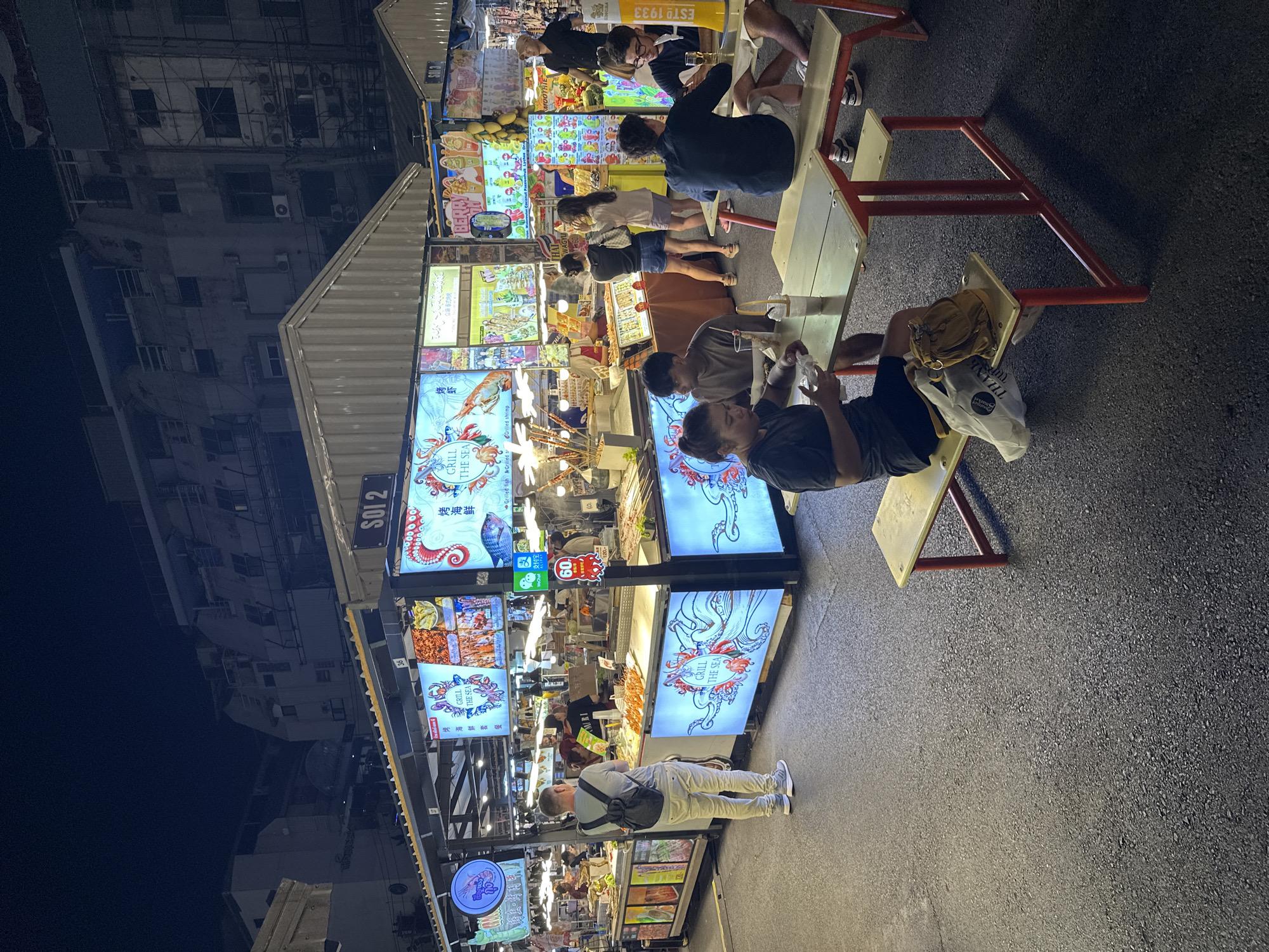 The image depicts a street food market at night, likely in Bangkok, Thailand. A row of food stalls is brightly lit, showcasing various seafood dishes and fruit smoothies. People are milling around, some seated at picnic tables, eating. In the foreground, a person sits on a bench, using a phone. Signs advertising "Grill the Sea" and "SOI 2" are visible. The architecture suggests an urban environment.