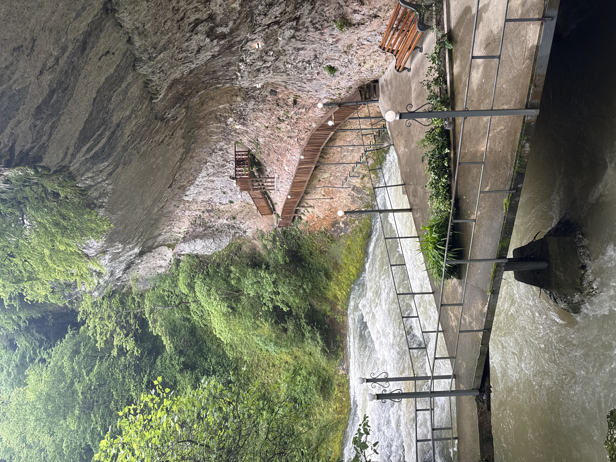 The image shows a view from a constructed pathway over a rushing river in Tsabal, Abkhazia. The pathway features metal railings and is partially supported by a concrete structure that extends over the river. A bench is placed near the edge of the pathway. A wooden staircase winds up a cliff face with light fixtures placed along the path. The river is turbulent and brown, reflecting the light from the sky. The surrounding landscape is lush with green vegetation and a large rock face, partially covered in foliage.