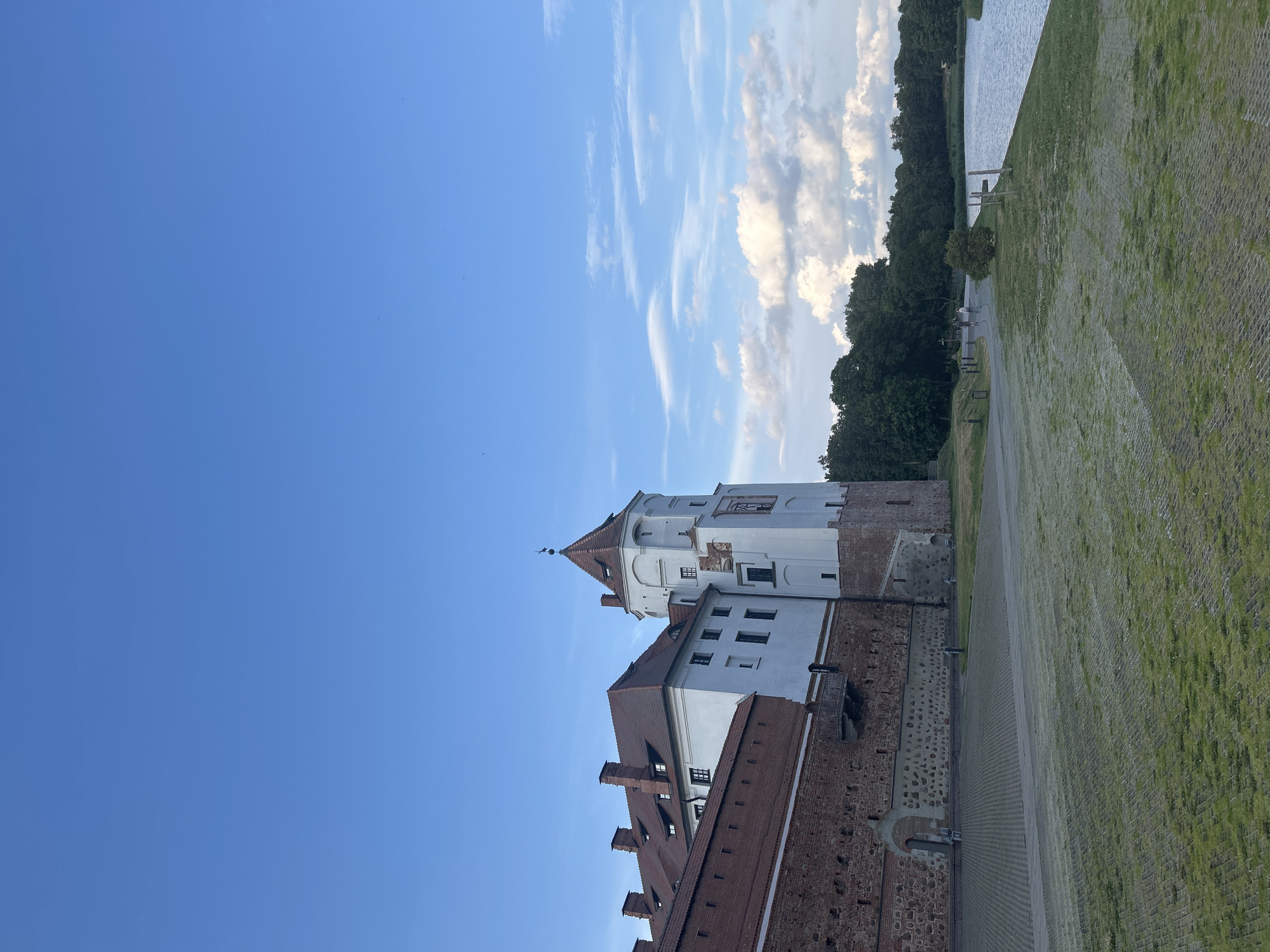 The image shows the Mir Castle Complex in the village of Mir, Grodno Region, Belarus, under a partly cloudy blue sky. The castle, a UNESCO World Heritage site, features a prominent white tower with a red, conical roof and a darker brick wall extending to the left, topped with red-tiled roofs. The castle is situated next to a calm body of water, likely a moat or lake, with a line of trees forming the background on the opposite bank. A paved area and a grassy expanse are in the foreground. Several indistinct figures are visible walking along the path by the water and near the castle entrance.