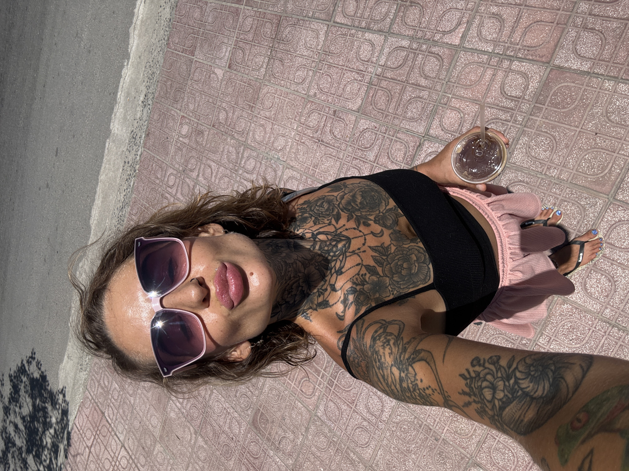 A person with extensive tattoos on their neck, chest, and arm is taking a selfie outdoors. They are wearing pink sunglasses, a black top, and a pink skirt. They are holding a clear plastic cup with a straw containing a dark liquid. The person is standing on a patterned tiled sidewalk next to a paved road. The scene suggests a sunny day. The location is described as Bac Nha Trang Ward, Vietnam.