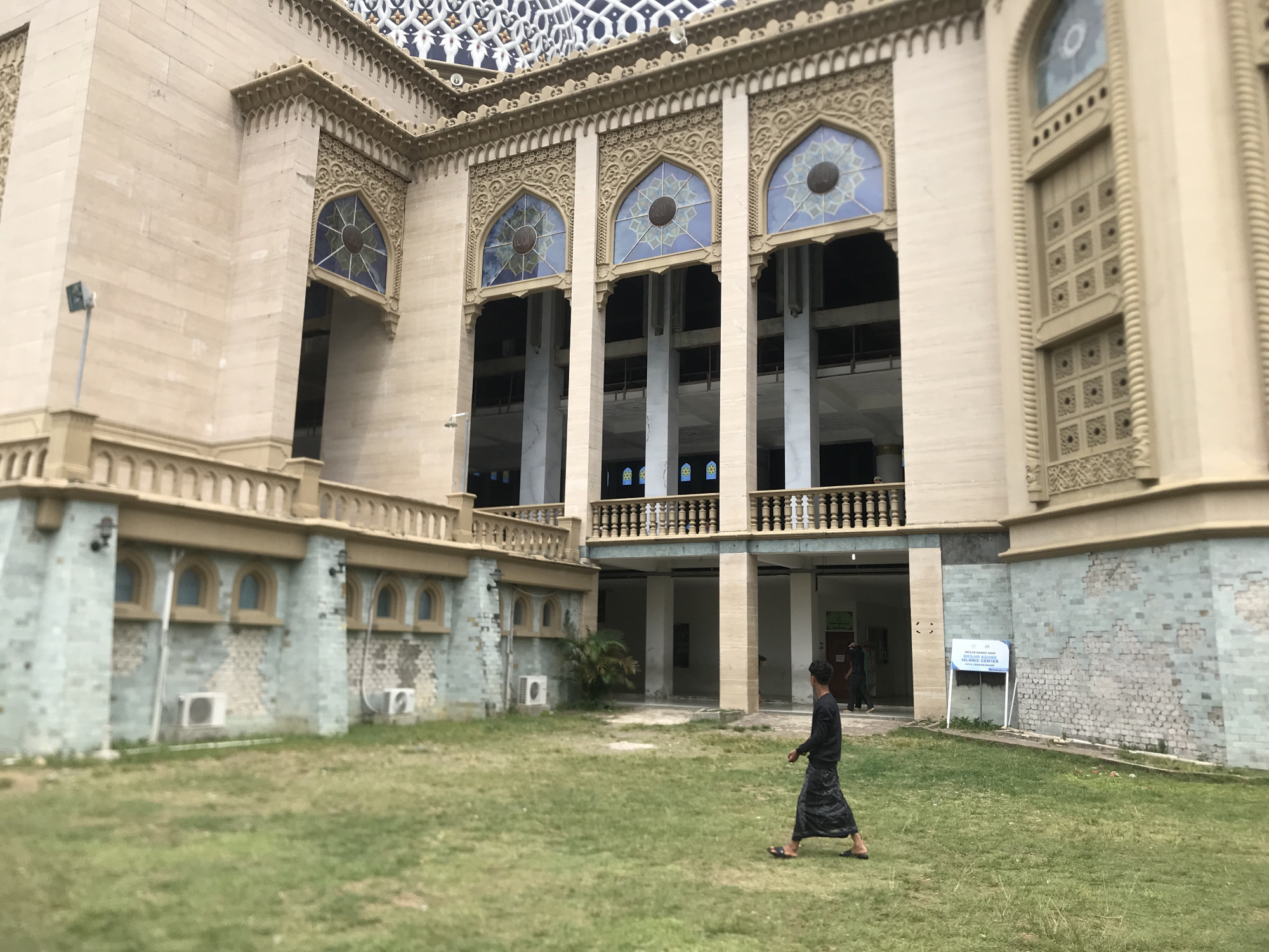 The image shows the exterior of the Islamic Center Mosque in Lhokseumawe, Indonesia. The mosque features a tan-colored facade with intricate architectural details, including decorative carvings around arched windows. Several tall, slender columns support an upper level with a balcony. The windows are adorned with stained glass featuring a circular motif. In the foreground, a grassy courtyard is visible, with a person in traditional dark clothing walking across it. In the background, the mosque's entrance is partially visible, and there is a blue and white sign on a stand. The sky is overcast, suggesting a cloudy day.