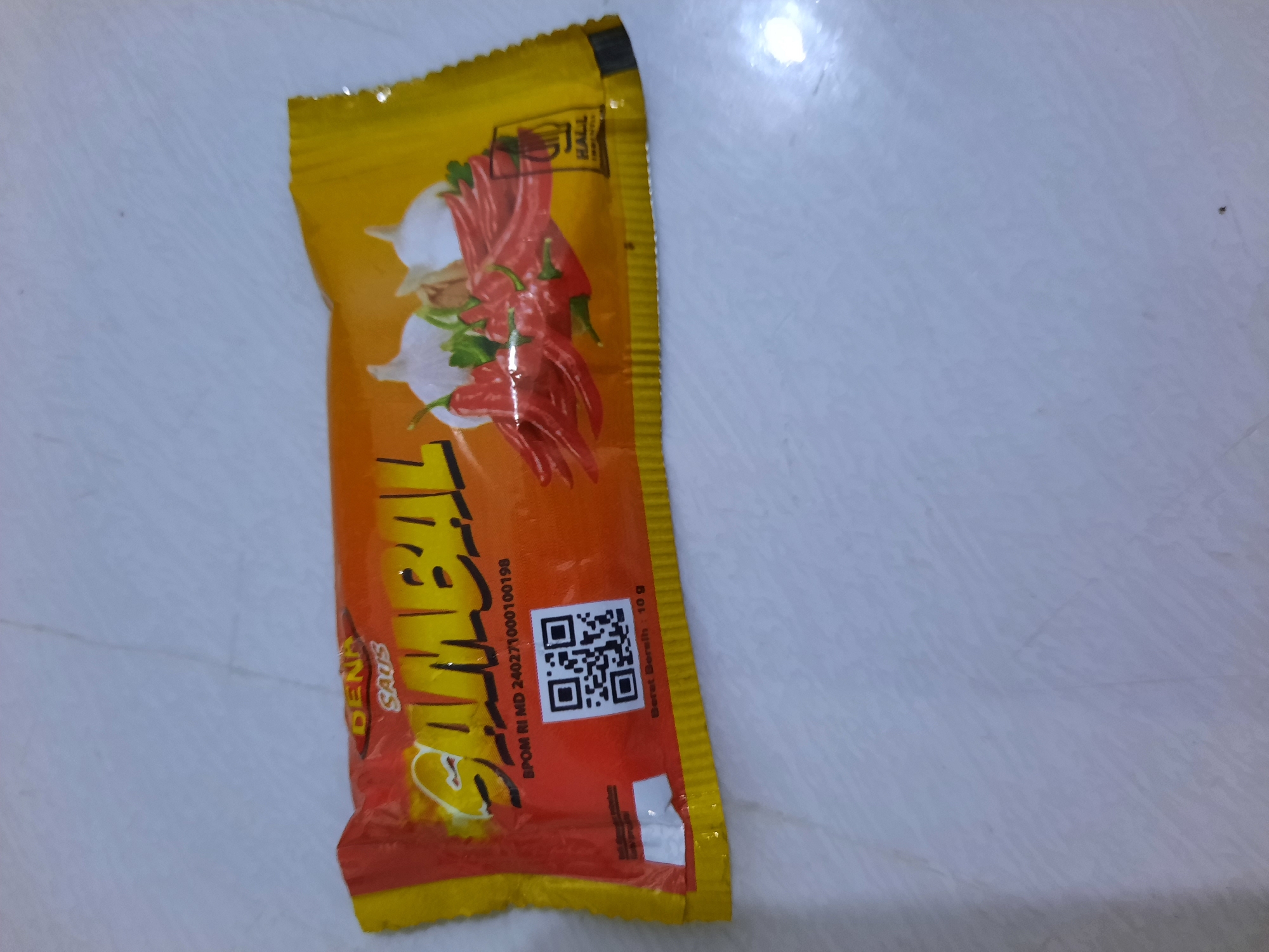 The image shows a package of "DENA SAMBAL" chili sauce on a light-colored, textured surface that resembles a floor or table. The packaging is primarily orange and yellow, with red chili peppers and white garlic depicted in the design. There is a QR code on the front of the package. The lighting suggests an indoor environment. There is no indication of the city of Lhokseumawe, Indonesia, or any of its features in the image.