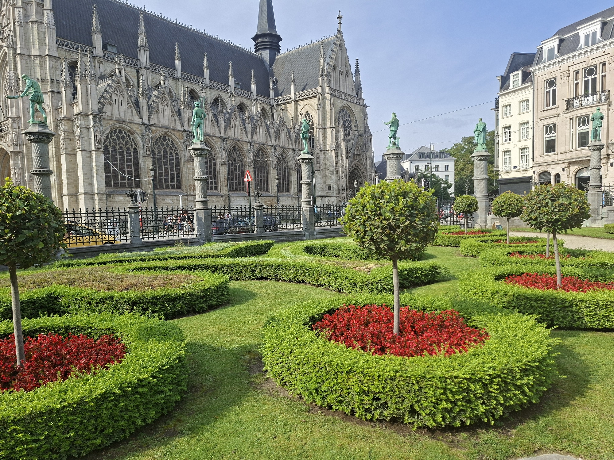 The image depicts a manicured garden in front of a grand gothic-style building in Brussels, Belgium. The building, likely a cathedral or large church, features ornate stonework, pointed arches, and tall spires. Several statues of men in historical attire are mounted on pedestals along the front of the building. The garden itself is laid out in formal patterns with neatly trimmed green hedges forming curved beds, interspersed with vibrant red flowers. Small trees with rounded canopies are strategically placed within the garden. In the background, beyond the garden and building, are other city buildings and a clear blue sky. The scene suggests a well-maintained public space, likely a tourist attraction or historical site.