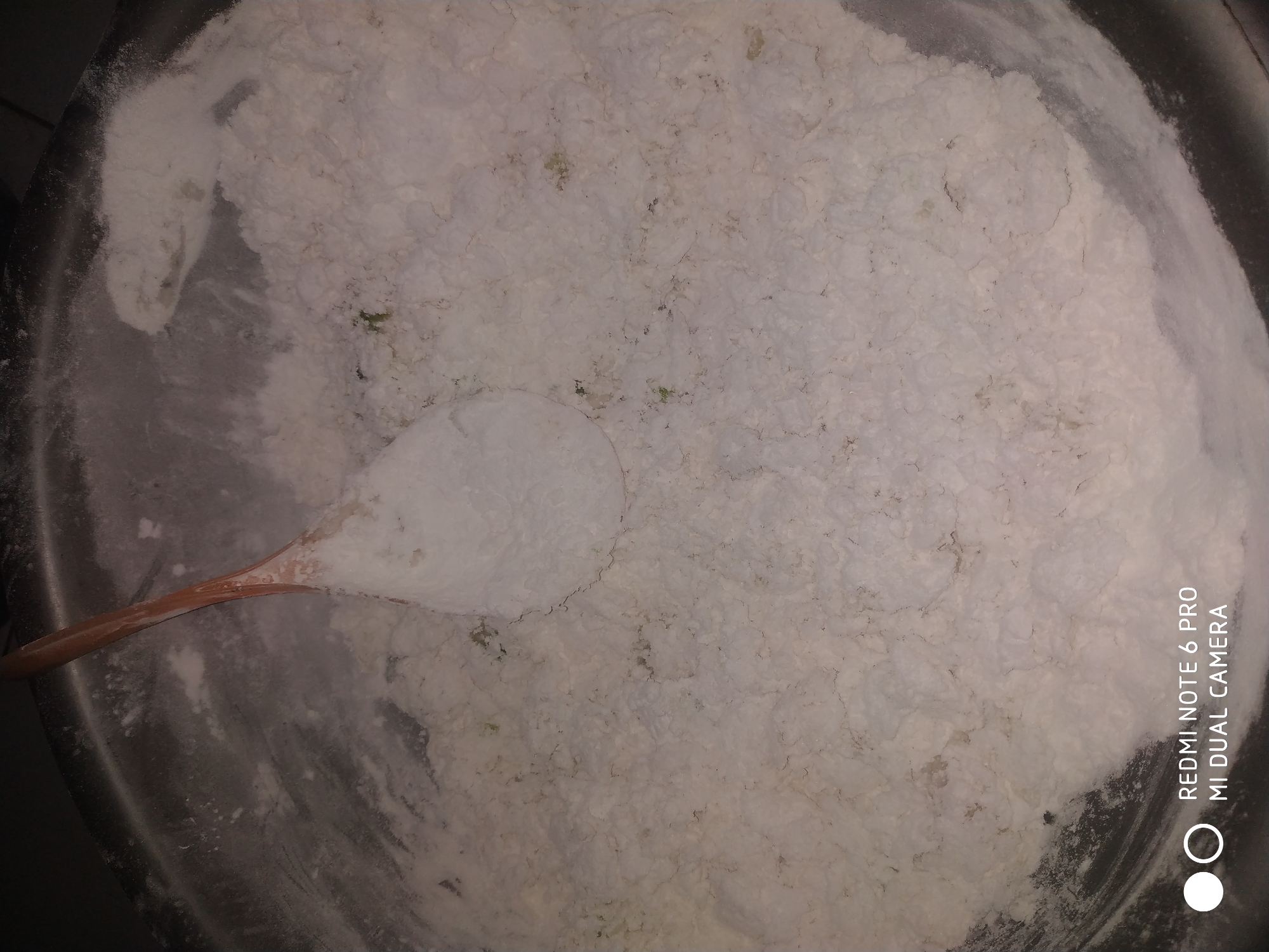 A wooden spoon is seen in a metal bowl filled with a white powdery substance, possibly flour or a similar ingredient. The substance appears to be textured, with some clumping and small green specks visible, suggesting it might be a mix. The background is dark, with the metallic sheen of the bowl partially visible. The bottom right corner of the image displays a watermark indicating it was taken with a "REDMI NOTE 6 PRO MI DUAL CAMERA." The scene is indoors, likely a kitchen environment in Badas, Indonesia.