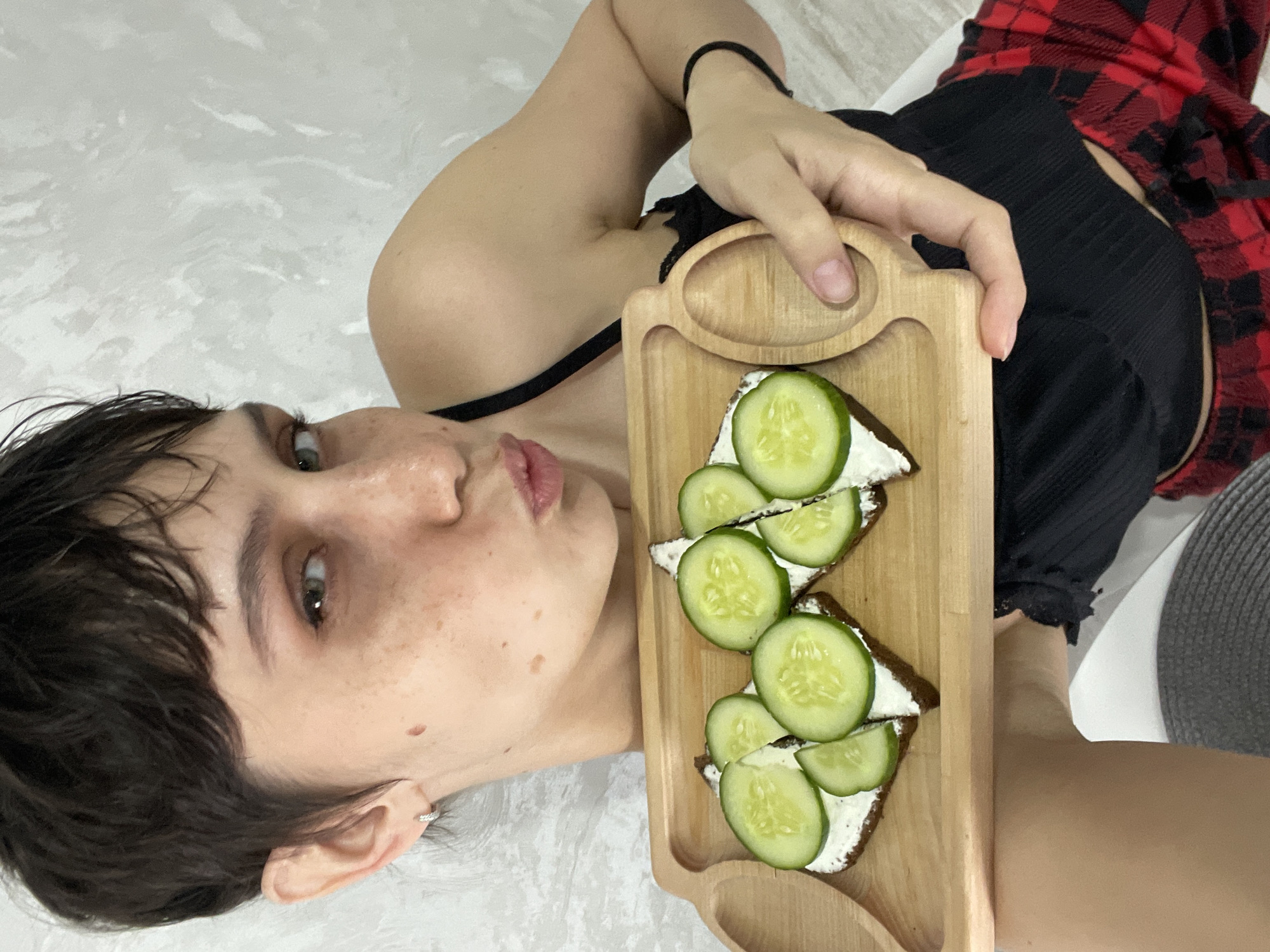 A woman is holding a wooden tray with three open-faced sandwiches. The sandwiches consist of dark bread, a white spread, and cucumber slices. She has short dark hair, fair skin with freckles, and is wearing a black tank top and a red and black plaid garment. The background is a textured white surface. The city of Izhevsk, Russia is not discernible from the image.