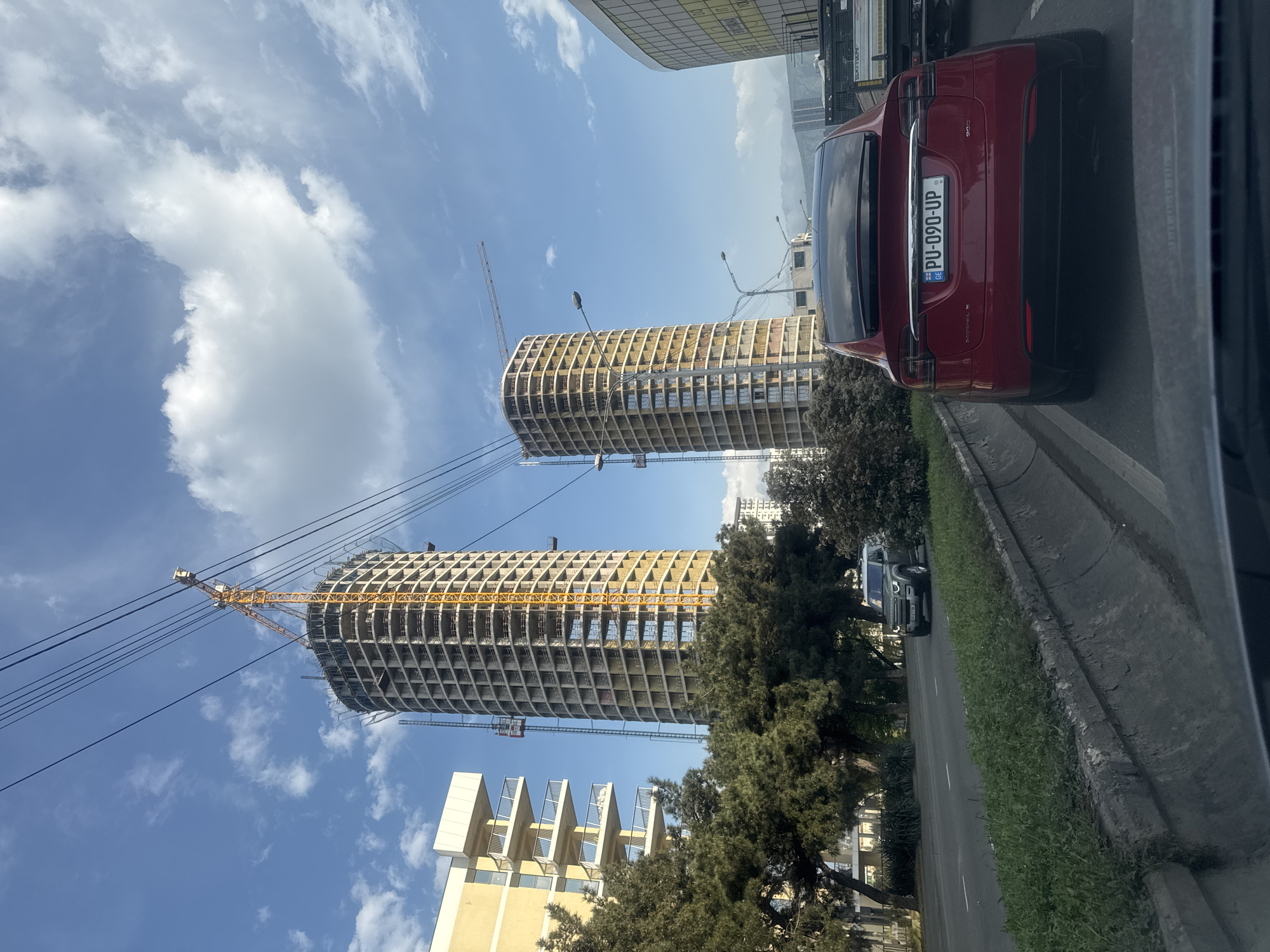 The image shows a view from a car in Tbilisi, Georgia. Two cylindrical buildings under construction dominate the left side of the frame, with yellow cranes attached. A lower, finished building with a yellow facade and balconies is also visible. The sky is blue with scattered clouds. On the right side of the frame, the rear of a red Tesla car is prominent, with the license plate "PU-090-UP," indicating a Georgian registration. A road with a grassy divider is present. Another vehicle is visible in the distance through the trees. The setting appears to be a modern area of the city with ongoing construction.