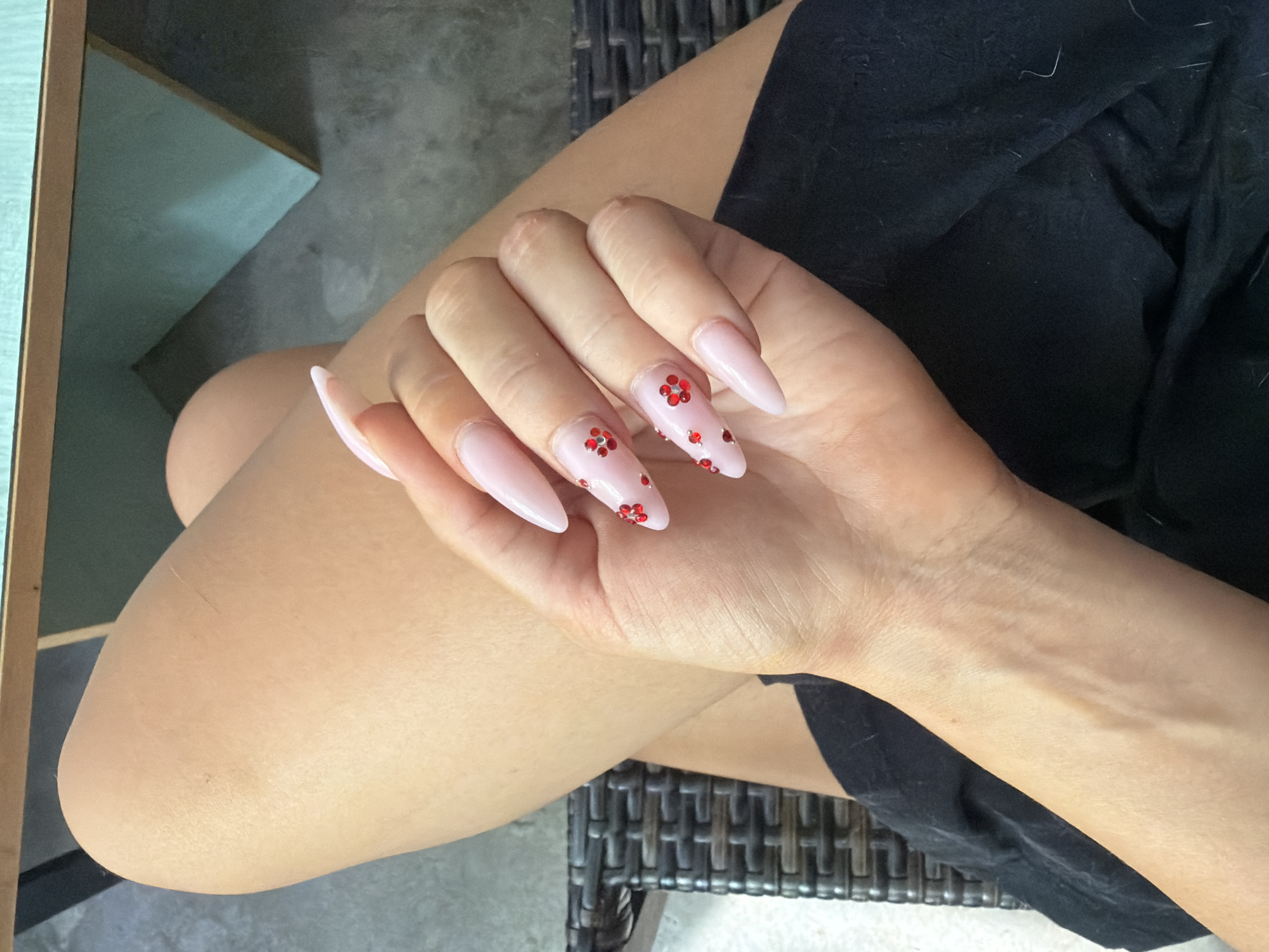 A close-up shot of a person's hand with light pink stiletto nails adorned with red glitter flower designs, resting on their thigh. The individual is wearing a black garment. The background includes dark wicker furniture and a light blue and brown table, suggesting an indoor setting or a shaded outdoor area in Prosena, Bulgaria.