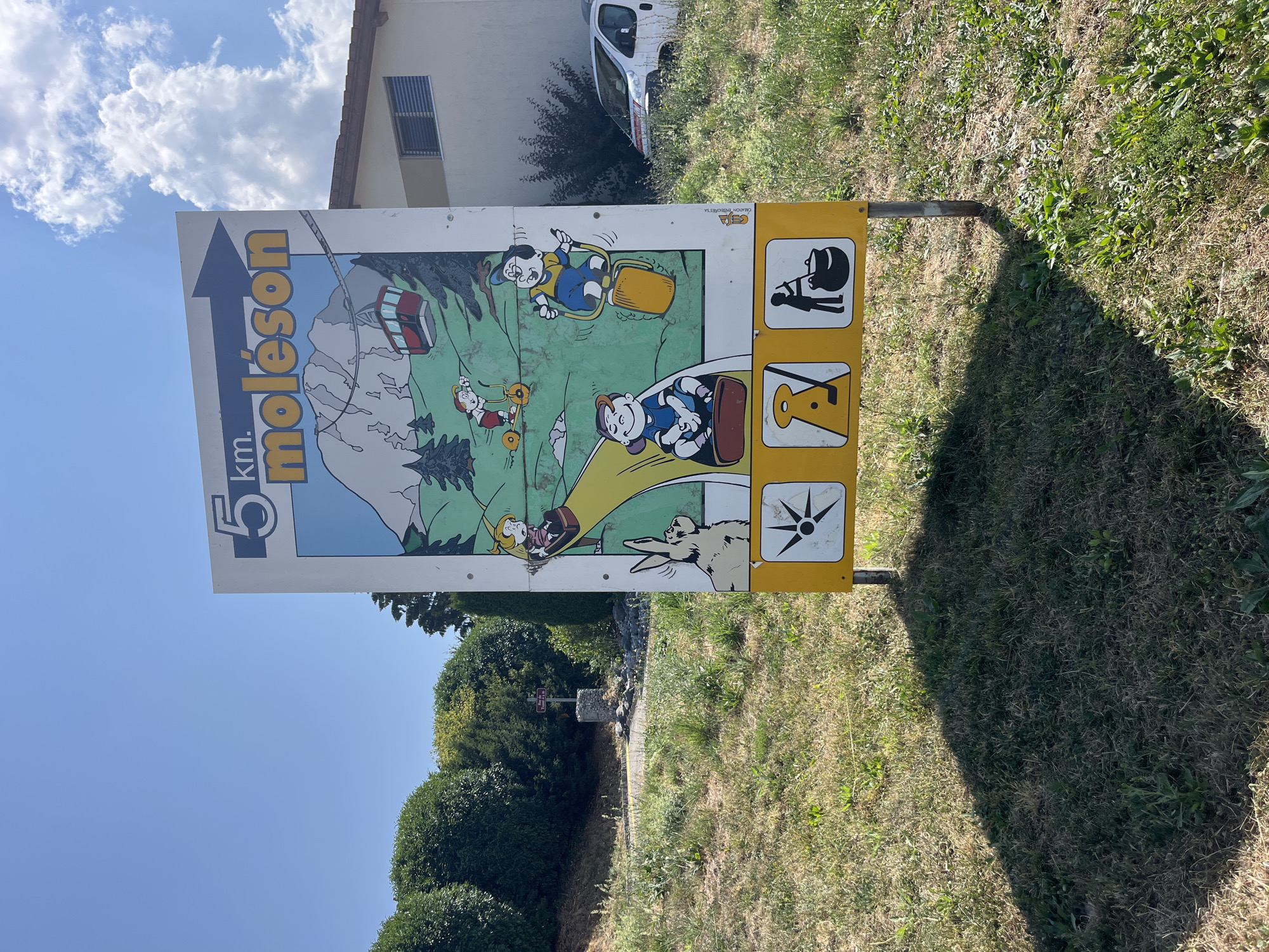 The image shows a tall, rectangular sign with a cartoon illustration on a white background. The sign indicates "5 km. moléson" with an arrow pointing to the right. The illustration depicts a mountain scene with a cable car, trees, and several cartoon characters engaged in activities. At the bottom of the sign, there are three yellow squares with icons: a sunburst symbol, a figure holding a golf club, and a figure stirring a cauldron. The sign is mounted on two metal posts and stands in a grassy area. In the background, there is a building with a light-colored facade and a dark roof, and a white van parked behind some bushes. The sky is blue with some scattered white clouds. The location is identified as Pringy, Switzerland.