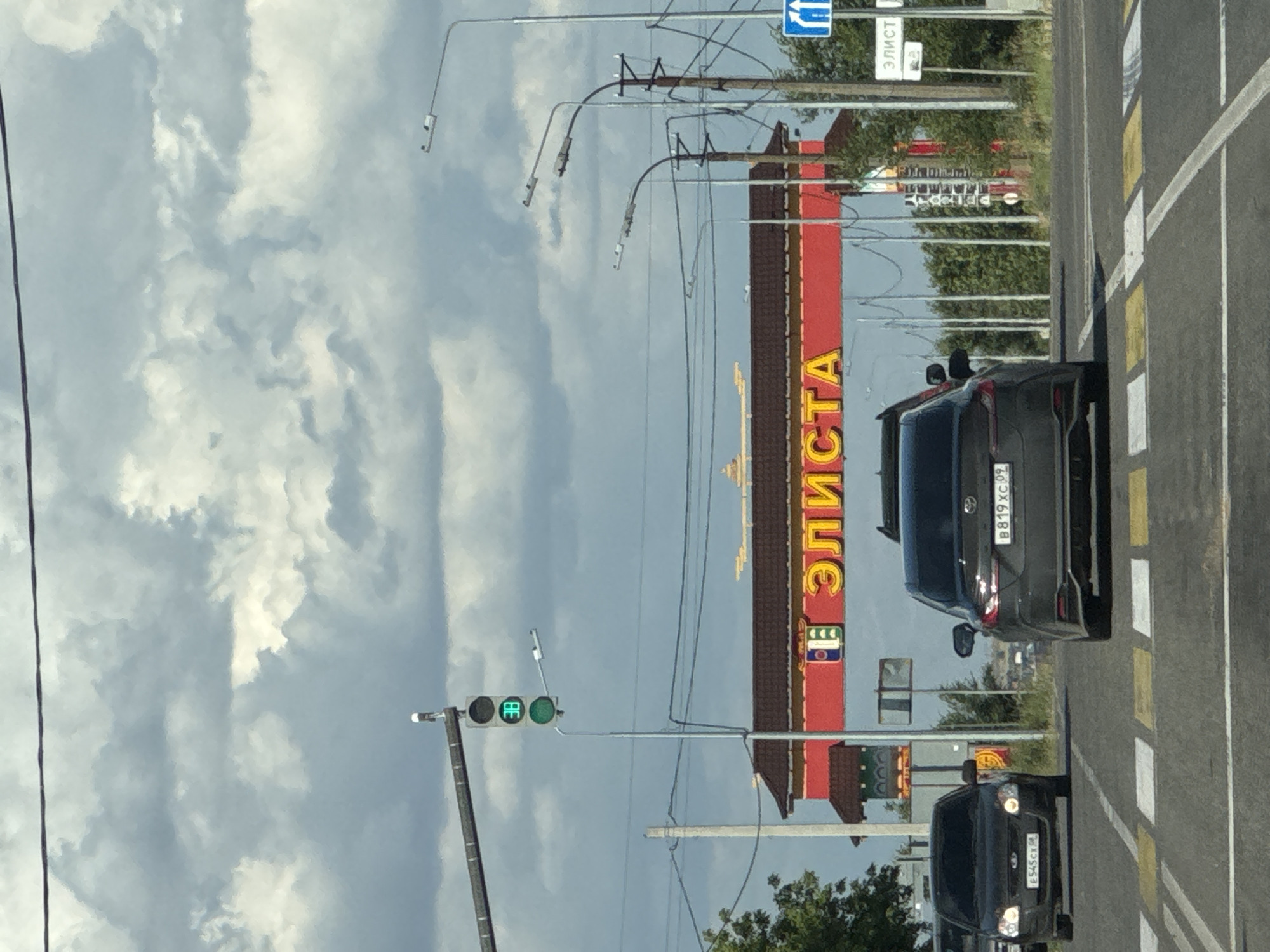 The image captures a street scene in Russia. The perspective is from a vehicle moving along a road, indicated by the presence of lane markings and other cars. The prominent subject is a large, red building with yellow lettering that reads "ЭЛИСТА." The building appears to be a commercial establishment, possibly a store or a business complex.
Several cars are visible on the road. One black car is parked in the foreground, and another black car is in the lane ahead, with its license plate partially visible as "B819XC 09". Further down the road, another black car with license plate "E545CH" is approaching. A traffic light showing a green signal is also present, suggesting the vehicles are at an intersection.
The sky is filled with dramatic, cloudy formations, suggesting a dynamic weather pattern, possibly overcast or transitioning. Streetlights and overhead wires are strung across the scene, typical of an urban or suburban environment. In the background, trees and other structures can be seen, contributing to the overall setting of a developed area. The time of day appears to be daytime, given the lighting conditions. No people are clearly discernible in the image. The overall impression is a snapshot of everyday life on a Russian road.