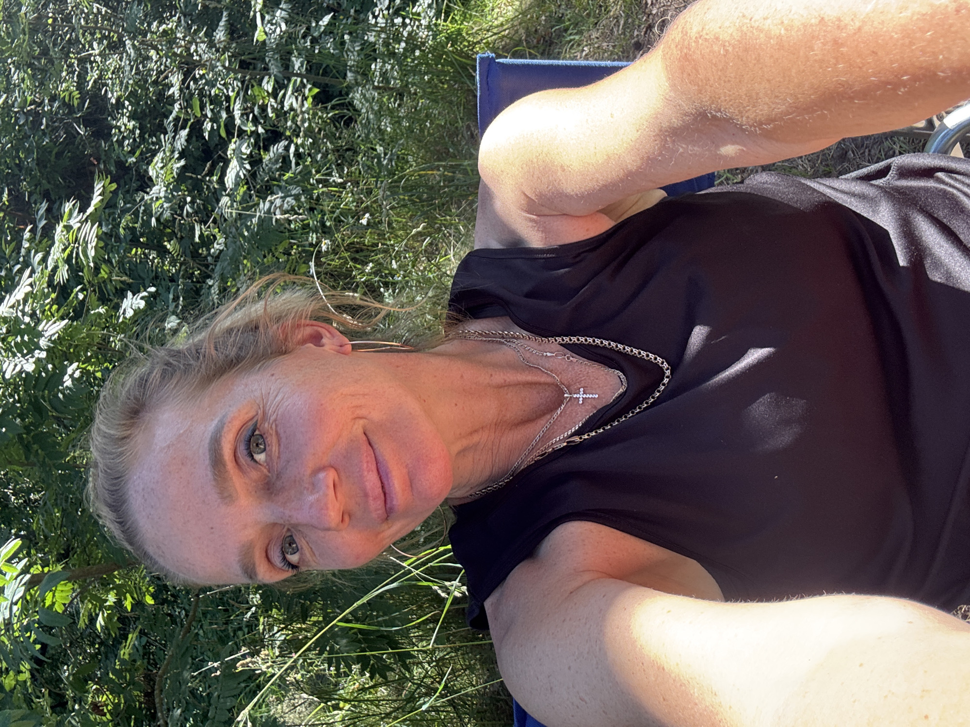 A woman with blonde hair pulled back, wearing a black tank top and a silver necklace with a cross pendant, smiles at the camera. She is sitting outdoors, with lush green foliage and trees visible in the background, suggesting a natural environment. The sunlight casts a warm glow on her face and arms. This scene is located in Zelenaya Roshcha, Russia, a place known for its natural beauty and recreational areas.