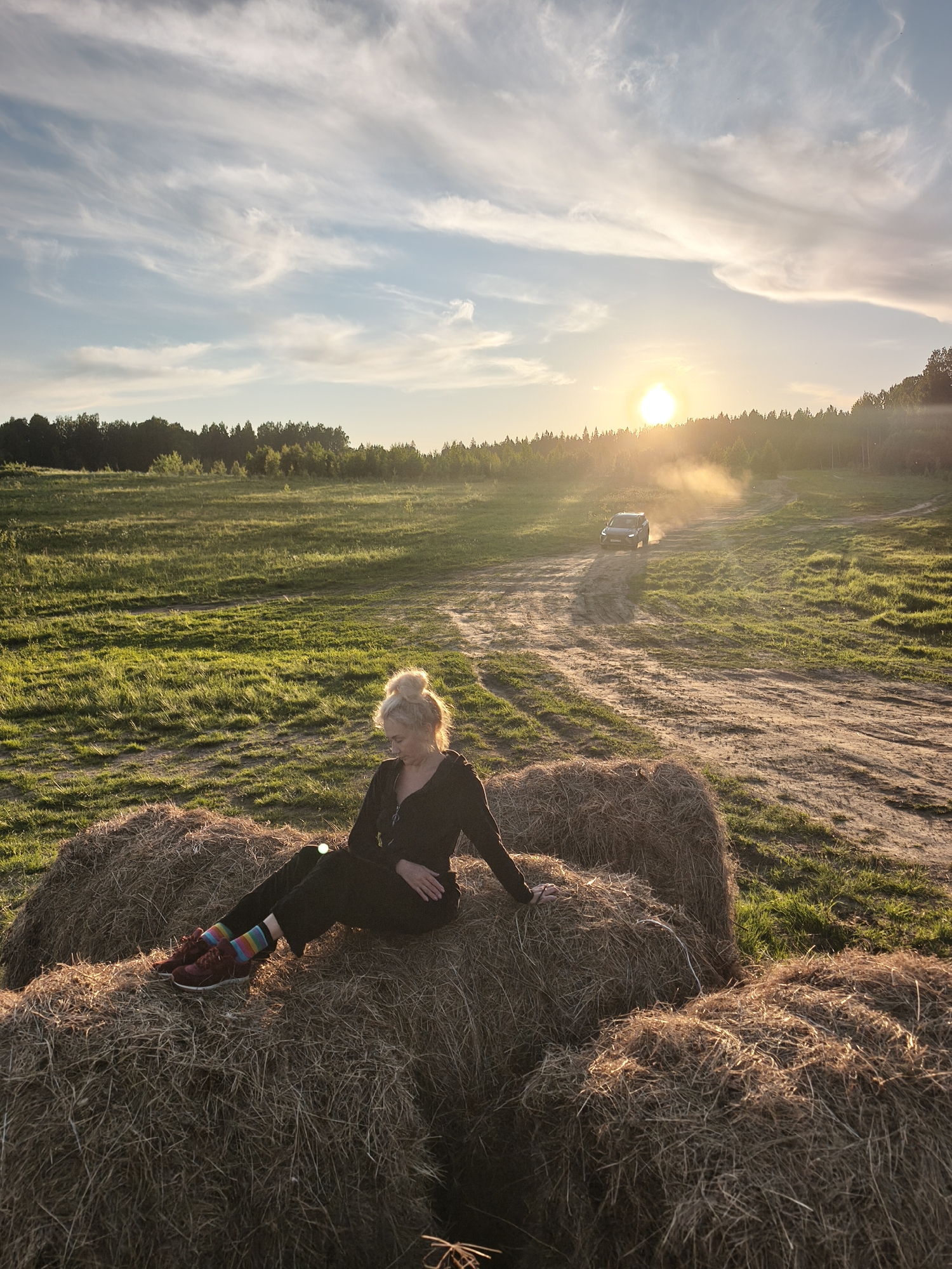 A wide shot captures a rural landscape at sunset in what appears to be S стеклянный, Russia. The warm, golden sun is low on the horizon, partially obscured by distant trees and casting a soft glow across the scene. The sky above features light blue hues with wispy, streaky white clouds.
In the foreground, a woman with blonde hair styled in a bun is seated comfortably on several large hay bales. She wears dark clothing and distinctive multi-colored, striped socks with dark red sneakers. Her head is tilted downwards, and she appears relaxed or contemplative, bathed in the warm sunset light.
Behind her, a vast green field stretches into the distance, bordered by a dense line of forest under the setting sun. A winding dirt road cuts through the field, and in the mid-ground, a dark-colored car is visible driving along this road, kicking up a trail of dust that is illuminated by the strong backlight of the sun. The overall atmosphere is peaceful and rustic, highlighted by the tranquil golden hour lighting. There is no visible text in the image.