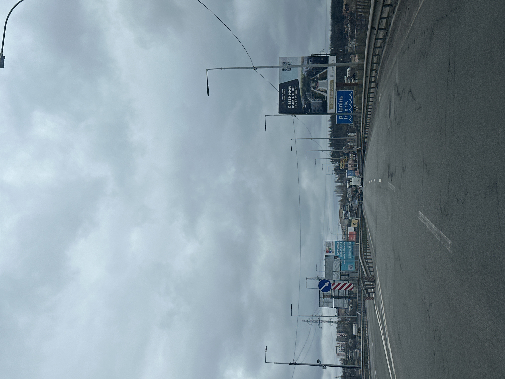 The image captures a street in Hostomel, Ukraine, under an overcast sky. The street is lined with billboards and streetlights. A sign indicates the direction and the name of the city "Irpin". The road surface is gray asphalt with faded white lane markings. Buildings and trees are visible in the background.
