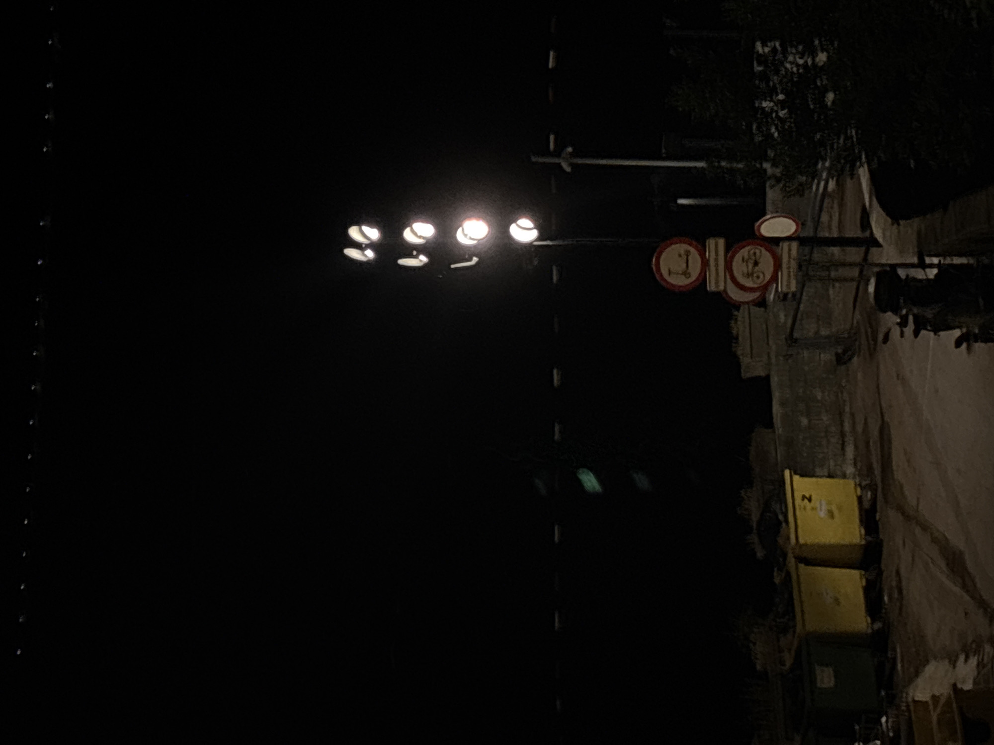The image captures a night scene in Herceg Novi, Montenegro. A row of streetlights illuminates a narrow road lined with buildings and trash bins. Several traffic signs are visible, indicating restrictions for scooters and bicycles. The overall environment appears dark and quiet.