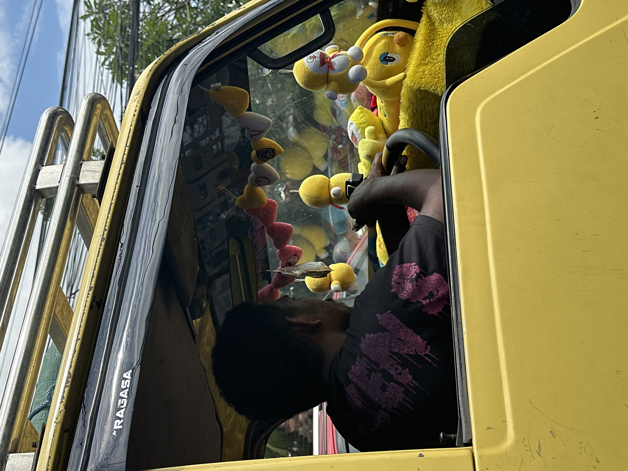 The image shows the interior of a yellow truck cab in Kuta Selatan, Indonesia. A man with short black hair is seated behind the steering wheel, wearing a black t-shirt with pink designs. The dashboard and front window are adorned with numerous yellow plush toys and hanging decorations. The truck's external roll cage and street scene are visible through the window. The word "RAGASA" is printed on the window's frame.