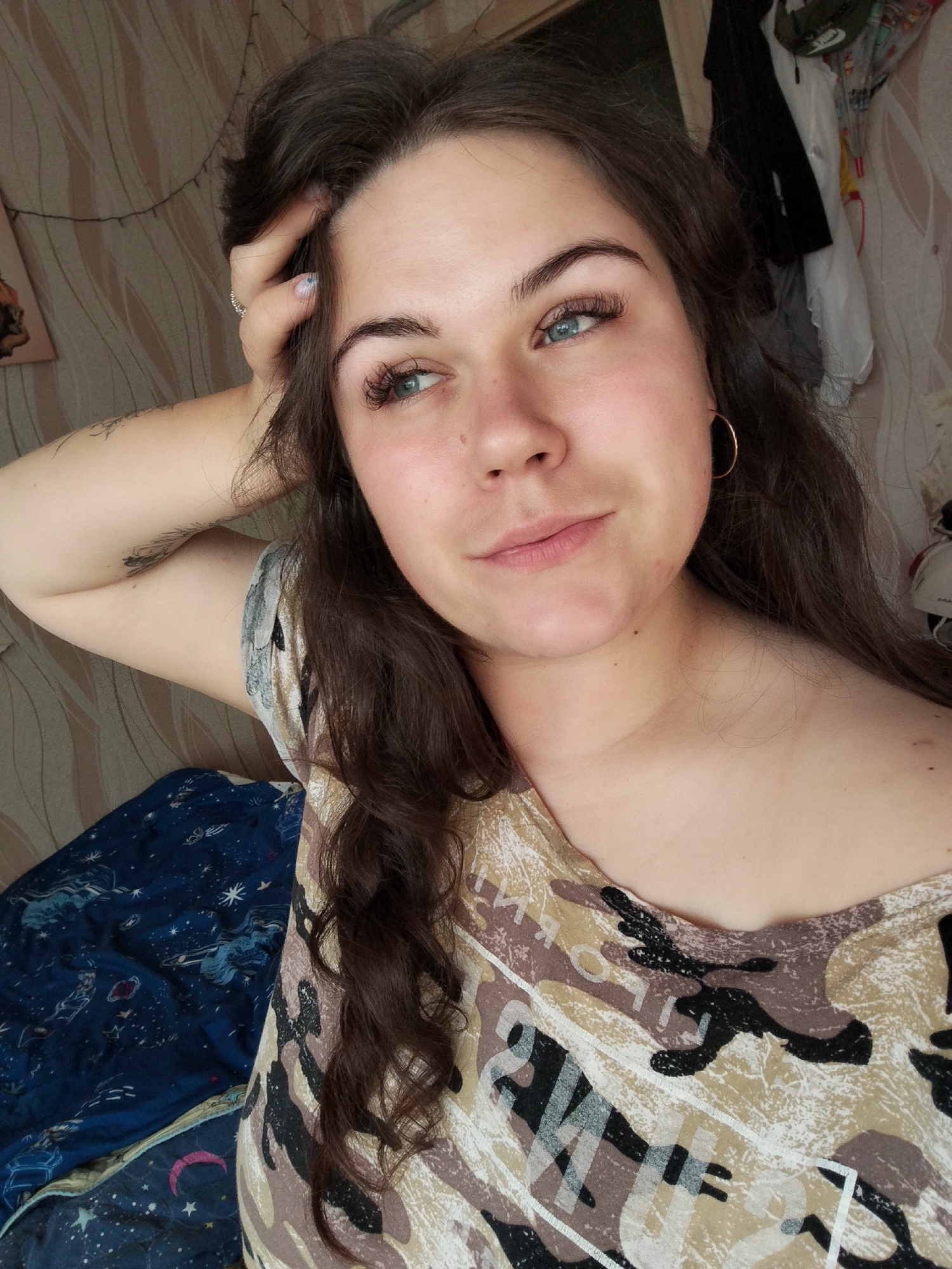 A young woman with long, dark brown, wavy hair and blue-green eyes is shown from the chest up, leaning her head on her right hand. She is wearing a camouflage-patterned shirt. Her left arm is bent behind her head, and her right arm is resting on her body. She has on hoop earrings. The background consists of a striped wall, a blue blanket with a celestial pattern, and some clothing hanging on the wall. The scene appears to be indoors, likely in a bedroom or living space. There is no specific indication of the city of Chaikovsky, Russia, in the image.