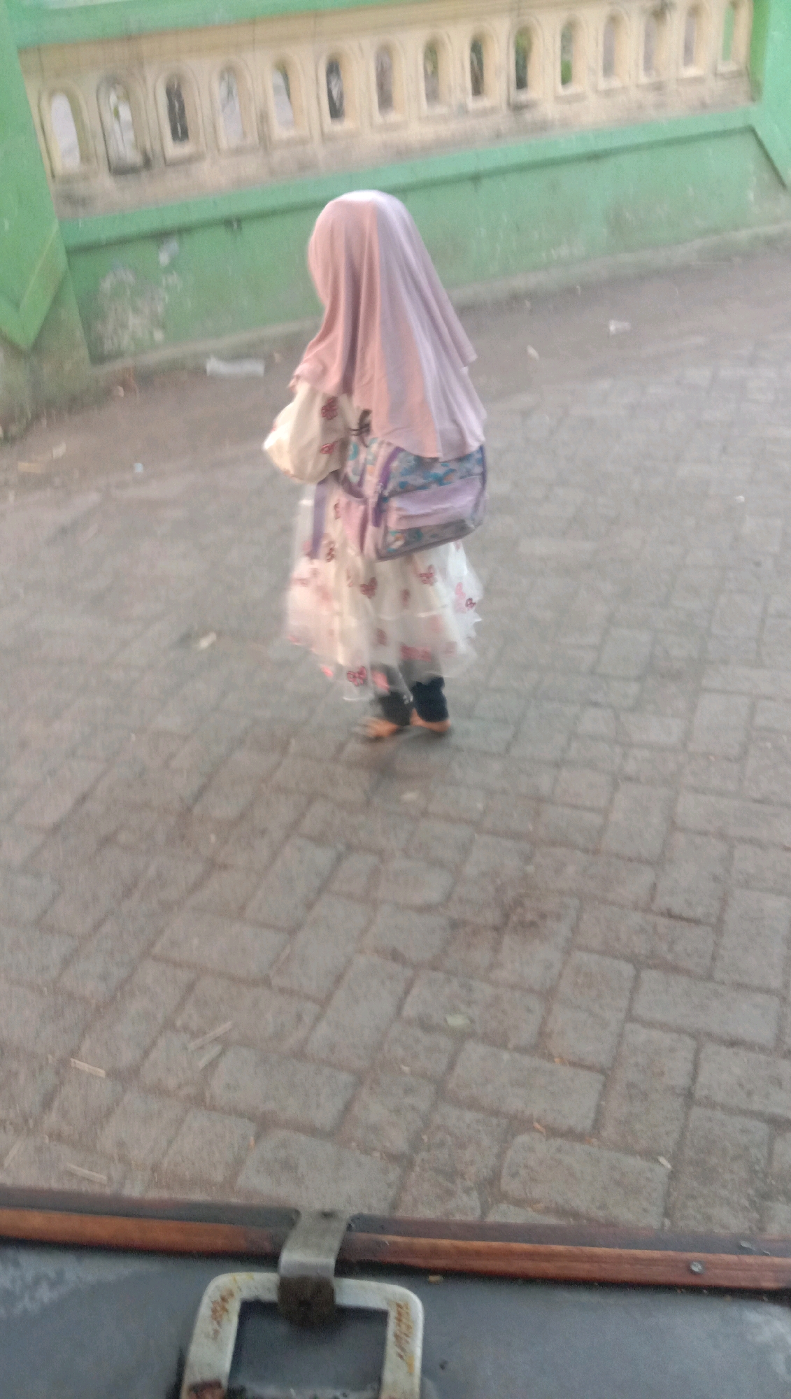 In Ngempit, Indonesia, a young child, possibly a girl, is seen from the back walking on a paved surface. She is wearing a light pink hijab, a white dress adorned with pink butterfly patterns, and dark shoes. A patterned bag, with shades of purple and blue, is slung over her shoulder. The background features a green wall with a decorative white railing featuring arched openings. The ground is composed of grey rectangular pavers, and the lighting suggests it is daytime. The foreground shows a dark surface with a metal latch, possibly part of a vehicle or furniture.