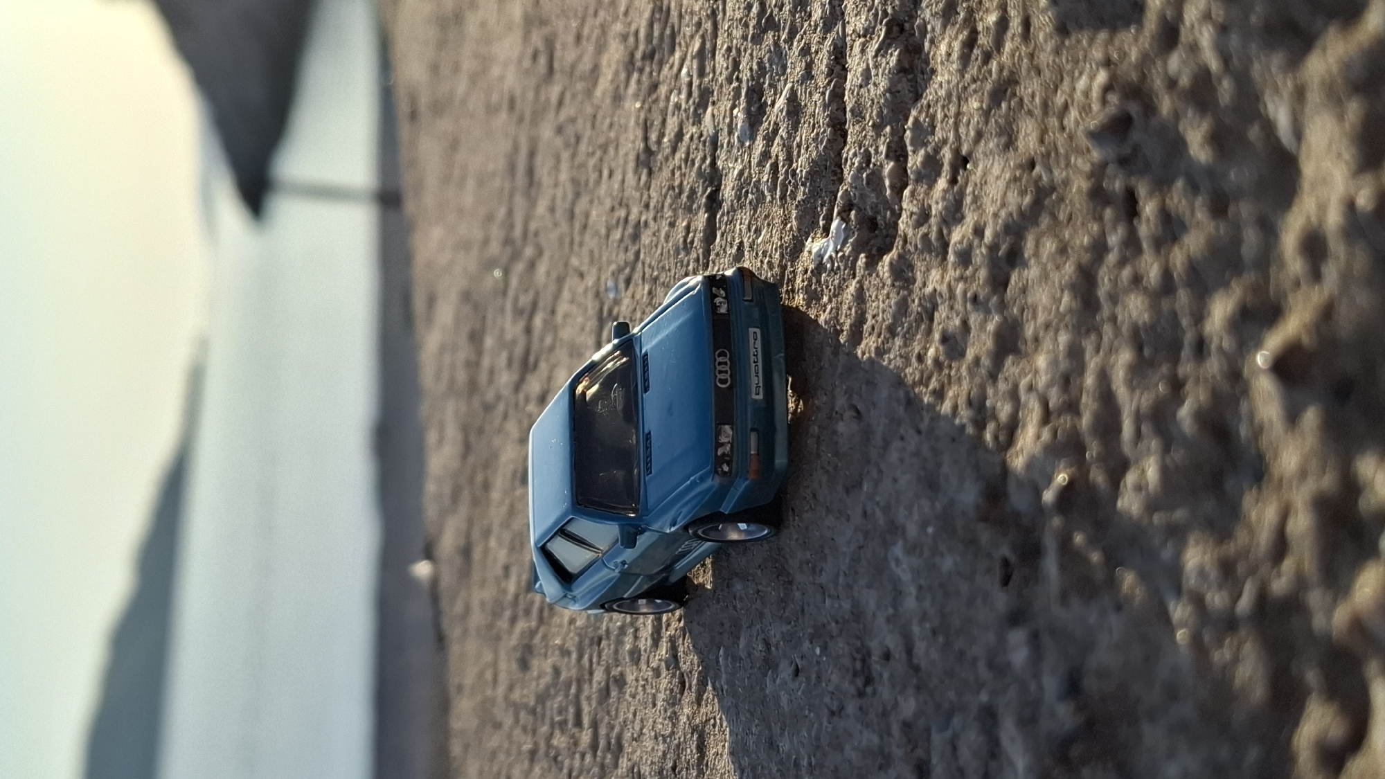 The image, captured in Boreti, Montenegro, showcases a detailed, teal-colored model car, specifically an Audi Quattro, placed on a rough, textured surface that appears to be concrete or stone. The photograph is oriented vertically, making the ground appear as a wall or steep incline.
Bright, direct sunlight illuminates the scene, evidenced by the distinct shadows cast by the car and the prominent texture on the ground, suggesting a clear and sunny day, likely mid-day or late afternoon.
The toy car features the iconic Audi rings logo on its front grille, with the word "quattro" clearly legible in white lettering beneath it. On what would be the license plate area, "AUDI SPORT" is visible, along with alphanumeric characters that resemble "A K C 2 7 9". The car's wheels have silver-colored rims.
In the background, a large, light-colored, and heavily blurred area extends, suggesting an outdoor environment with a distant structure, wall, or the sky. There are no people visible, and the car is stationary, presented as a still-life representation.