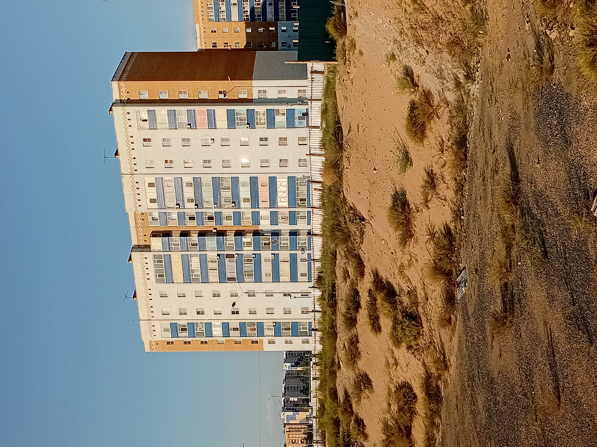 The image captures a residential apartment building in the city of Semey, Kazakhstan. The building is tall and rectangular, with a facade primarily of white and blue. The right side of the building is a contrasting orange-brown brick. Rows of windows, some with blue trim, are visible across the building's many floors. Decorative elements, such as a patterned frieze, are present near the roofline.
In the foreground, there is a sandy embankment with sparse, dry vegetation. A dark metal fence runs along the base of the embankment. Beyond the main building, other apartment buildings of similar architectural style can be seen in the distance. The sky is a clear, pale blue. The ground in the immediate foreground is a mix of gravel and dirt. The overall scene suggests an urban environment, possibly a developing or recently constructed area, with evidence of arid or semi-arid landscaping.