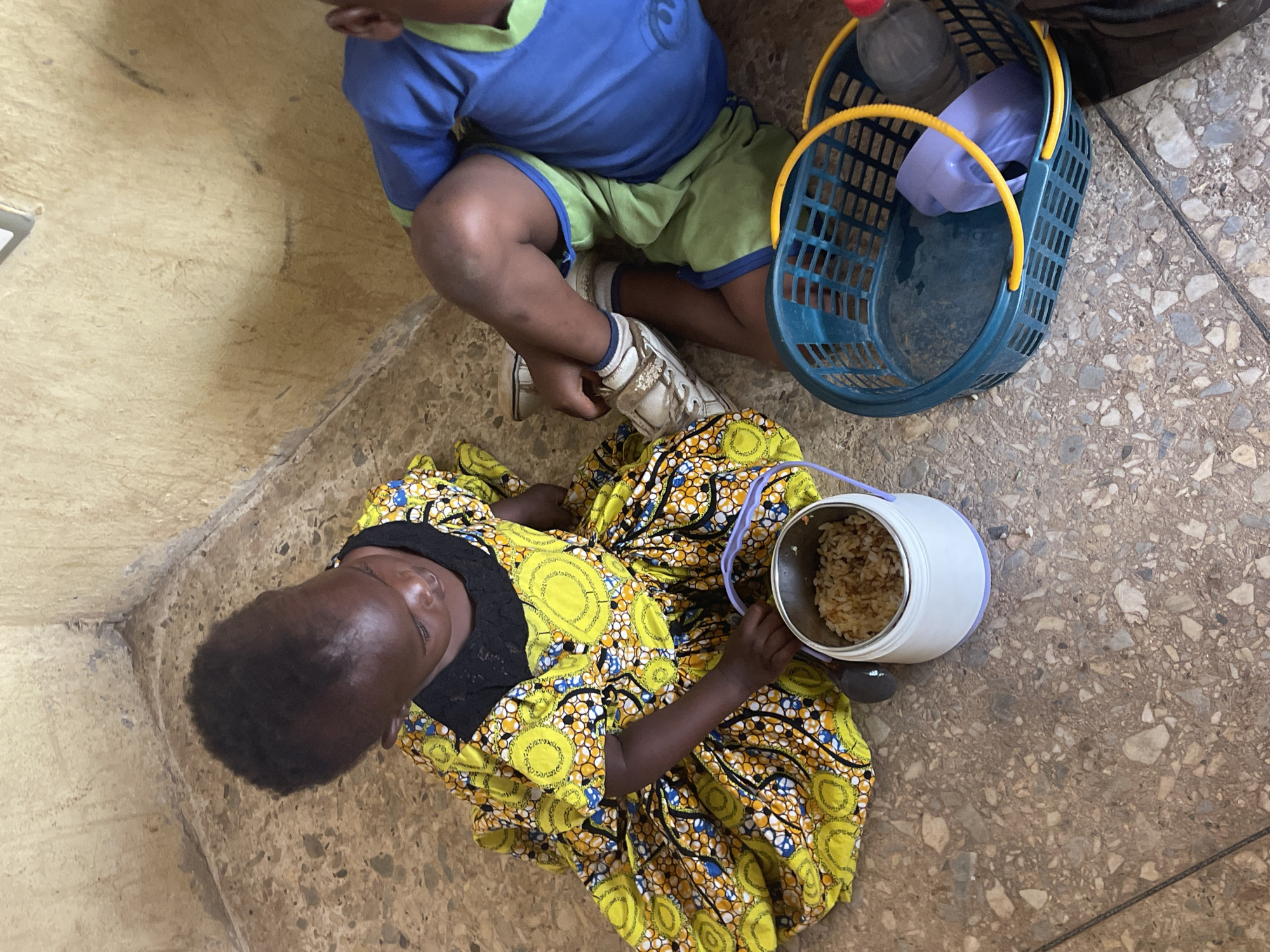 An overhead shot shows two individuals, an adult and a child, seated on a speckled concrete floor next to a plain, light-colored wall. The adult, with dark skin and short dark hair, is on the left, wearing a vibrant yellow, blue, and black patterned dress. She holds an open white and silver insulated food container with a purple handle, which contains a meal of rice. A dark-colored spoon is visible next to the container.
The child, also with dark skin, is seated to the right of the adult, facing away from the camera. He wears a blue t-shirt with a light green collar and short sleeves, paired with light green shorts that have blue trim. He has white sneakers on his feet.
Next to the child is a small, rectangular blue plastic basket with yellow handles. Inside the basket are a clear plastic water bottle and a light purple plastic cup or bowl. The scene depicts a moment of rest or mealtime. The setting appears to be indoors, likely a simple building in Akwanga, Nigeria. No visible text is present in the image.