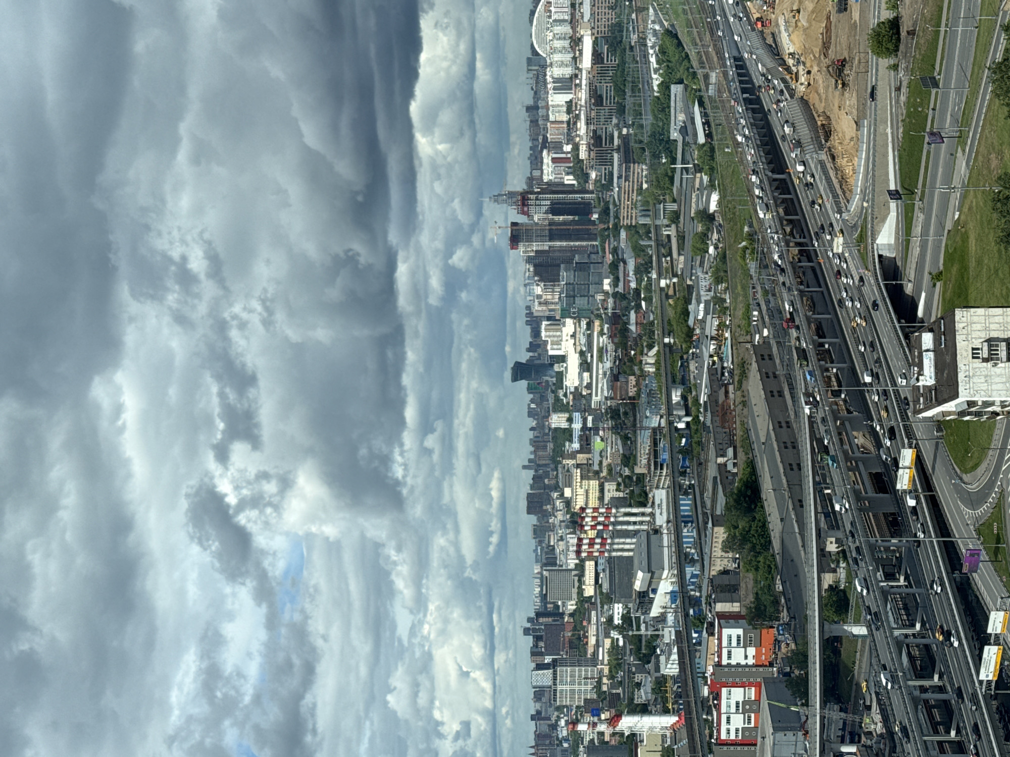 The image depicts a high-angle view of Khimki, Russia, on a cloudy day. The left side of the image is dominated by a sky filled with layers of gray and white clouds. The right side reveals the cityscape, featuring a mix of residential and commercial buildings of varying heights, interspersed with greenery. Several roadways and railways stretch across the cityscape, exhibiting moderate to heavy traffic. In the upper portion of the cityscape, construction work is visible, with earthmoving equipment and exposed soil.