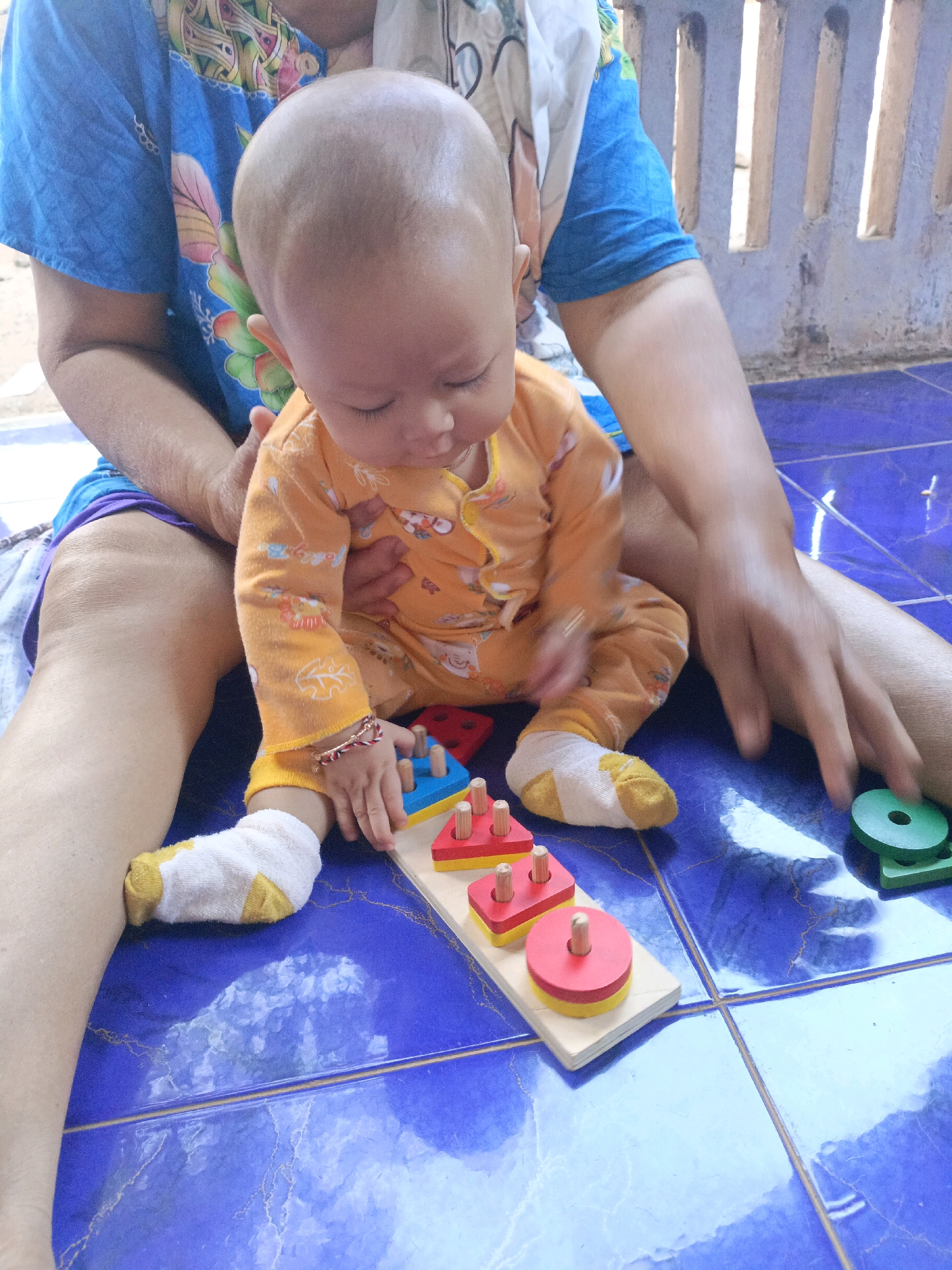 The image depicts a baby sitting on a blue tiled floor, held by an adult. The baby, wearing an orange outfit and white socks with yellow accents, is looking down at a wooden shape sorter toy. The toy features red and yellow geometric shapes, including circles and triangles, on wooden pegs. To the right of the toy, an adult's hand is interacting with green shape pieces. The adult is wearing a blue shirt with a colorful floral pattern. The environment appears to be outdoors, with a concrete wall in the background that has vertical openings. The scene is set in Jenu, Indonesia.