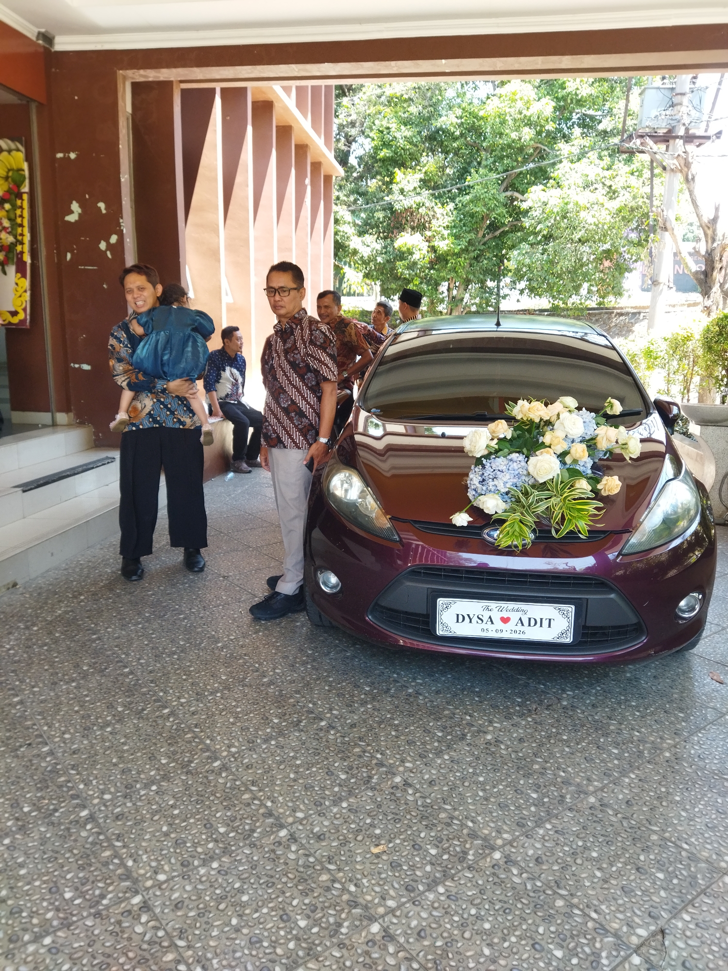 In Semarang, Indonesia, a burgundy Ford Fiesta, decorated with a bouquet of white and yellow roses and blue hydrangeas on its hood, is parked on a textured gray tiled surface. A wedding license plate reading "The Wedding DYSA ADIT 05.09.2026" is affixed to the front of the car. To the left of the car, a man wearing a batik shirt and black pants carries a young child dressed in a blue outfit. Beside him, a man in a similar batik shirt and light gray pants stands looking towards the car. Other individuals, some wearing batik and one in a black skullcap, are visible in the background, standing or sitting near a building with brown and beige architectural elements. Lush green trees and utility poles are visible beyond the building. The scene appears to be outdoors, likely in front of a building or event venue.