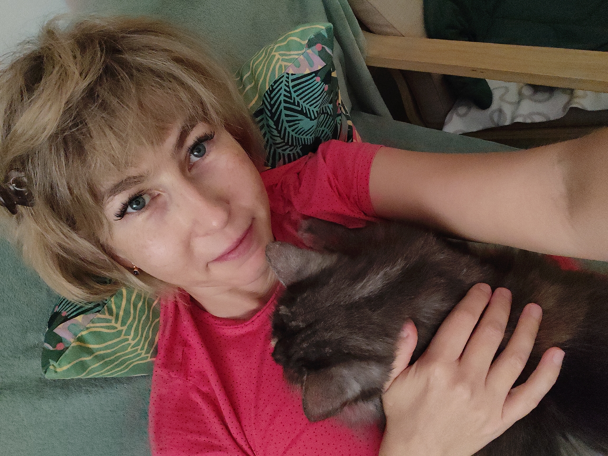 In a close-up, high-angle selfie taken in Bol'shoye Kozino, Russia, a woman with blonde hair and blue eyes is lying down, holding a dark gray cat. She is wearing a pink t-shirt with a subtle pattern. The woman's left arm is extended, holding the camera, while her right arm is wrapped around the cat. The cat is nestled against her chest, with its head resting on her shoulder. The background includes a green couch with a pillow featuring a tropical leaf pattern, and a glimpse of wooden furniture. The lighting is soft, suggesting an indoor setting.