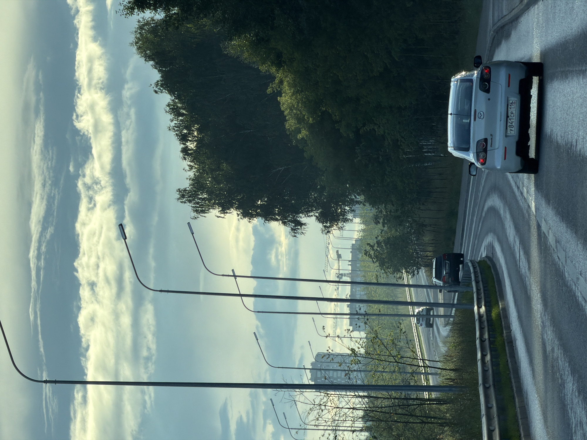 The image captures a view of a curved road in Kazan, Russia, on a cloudy day. The road, likely part of a highway or a major thoroughfare, is wet, possibly from recent rain, and has a light-colored barrier on the left side. Several streetlights line the left side of the road, with one prominent pole in the foreground.
In the background, there are buildings, including a tall apartment complex and construction cranes, indicating urban development. Lush green trees and vegetation flank the right side of the road, creating a contrast between the natural and man-made elements.
Three cars are visible on the road. In the foreground, a light-colored sedan with a Russian license plate reading "Y841CP 116" is driving away from the camera. Further ahead, two SUVs are also driving in the same direction. The sky is filled with dramatic, layered clouds, illuminated by sunlight filtering through, creating a dynamic atmosphere. The overall impression is that of a suburban or semi-urban environment during daytime.