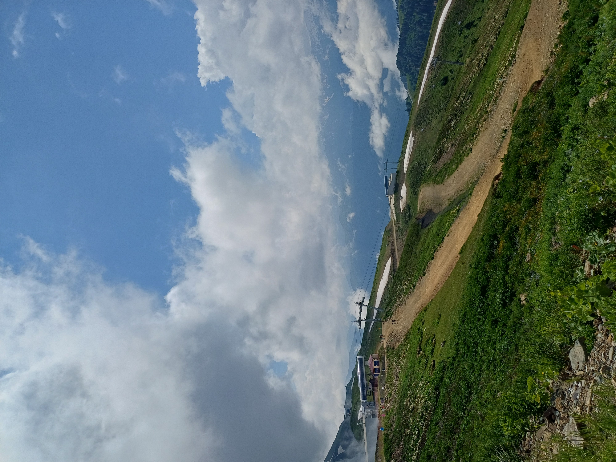 The image depicts a scenic view of a mountain landscape in Estosadok, Russia, likely during the summer months. The foreground is dominated by lush green vegetation and rocky terrain. A dirt path winds its way up the grassy slope, with patches of snow remaining in sheltered areas. In the mid-ground, a ski lift station is visible, with a cable car system extending across the mountainside. Several people are seen walking on the path and near the station. Further in the background, rolling hills and distant mountains can be observed, partially obscured by clouds. The sky above is a mixture of blue sky and fluffy white clouds.
