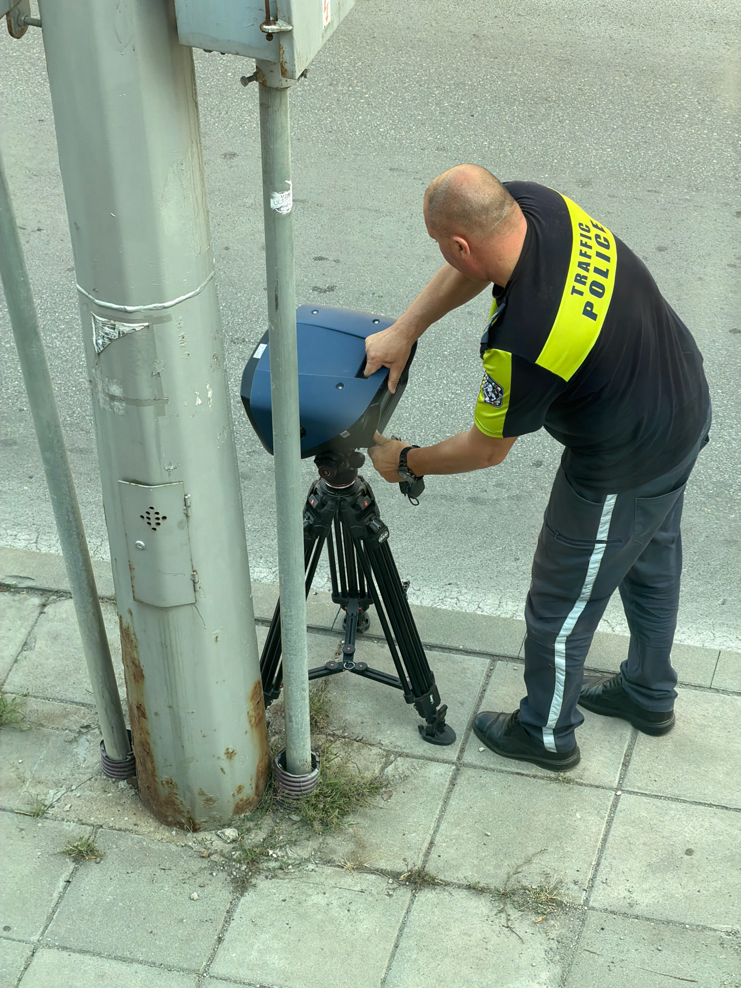 An overhead shot shows a bald male traffic police officer in Sofia, Bulgaria, adjusting a portable speed detection device mounted on a tripod. The officer wears a black t-shirt with a bright yellow band across the shoulders and chest, bearing the words "TRAFFIC POLICE" in yellow capital letters. He also wears dark grey pants with a reflective stripe down the side and black leather shoes. The speed detection device is a dark blue, aerodynamic unit. The scene takes place on a paved sidewalk next to a street. A thick, grey metal pole, possibly a lamppost or part of a traffic signal, stands to the left of the officer and the device. This pole shows signs of rust near its base, and has some stickers and a small metal cover with a grill pattern on it. The ground is a concrete sidewalk made of square tiles, with some patches of grass growing between the tiles, particularly near the base of the metal pole. The street in the background is asphalt.