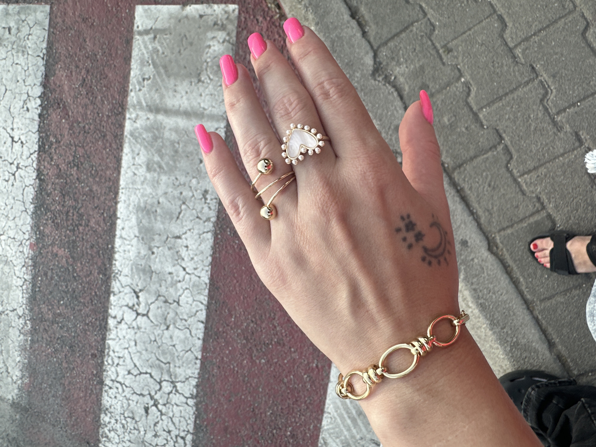 The image features a hand with pink manicured nails, adorned with gold jewelry including a heart-shaped ring, a spiral ring with gold beads, and a bracelet, positioned over a city crosswalk. A tattoo with a moon and stars is visible on the wrist. The crosswalk has a pattern of white and red stripes, while the background shows a gray brick sidewalk. A foot wearing a black sandal is visible in the background. The location is inferred to be in Budva, Montenegro, because the tags associated with this image include "Budva" and "Montenegro."