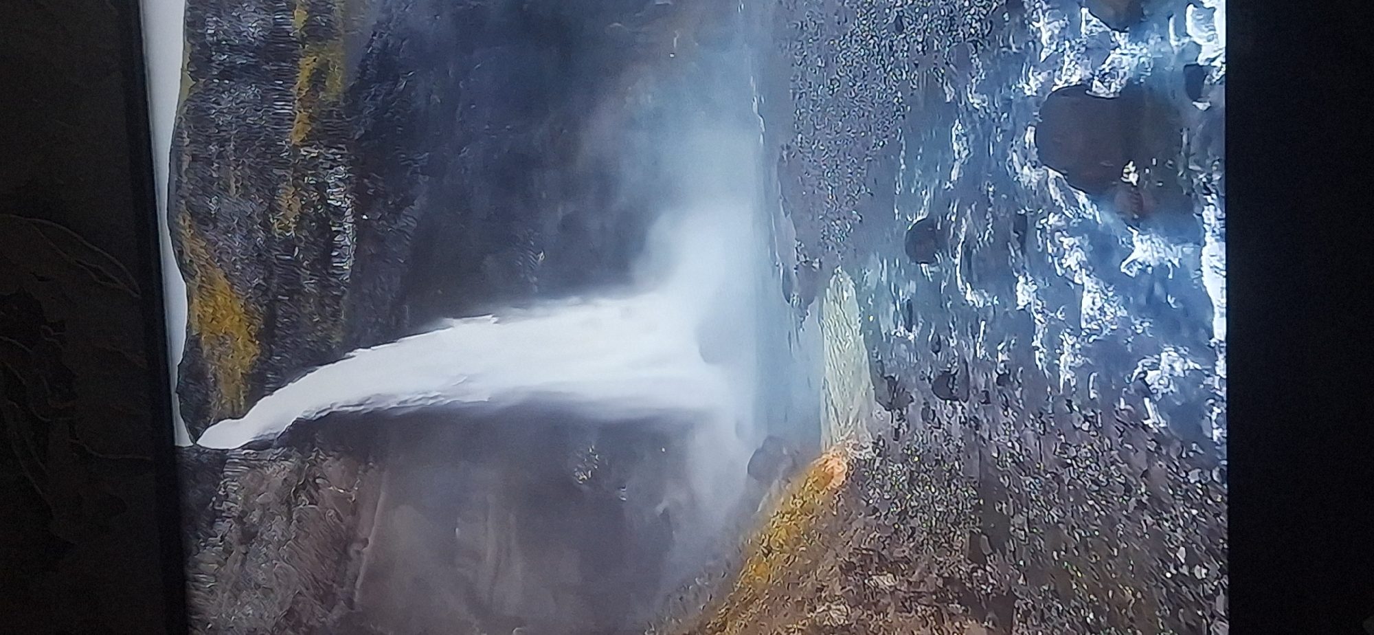 The image depicts a majestic waterfall cascading down a rocky cliff face. The water is white and frothy as it plunges into a pool below, creating a misty spray. The surrounding landscape is rugged and natural, with rocks and sparse vegetation on the slopes. The water in the riverbed is a murky greenish-blue, with many rocks visible, some submerged and others protruding from the water. The overall scene evokes a sense of raw, natural beauty. The image does not show any elements that would suggest a location in Bila Tserkva, Ukraine.