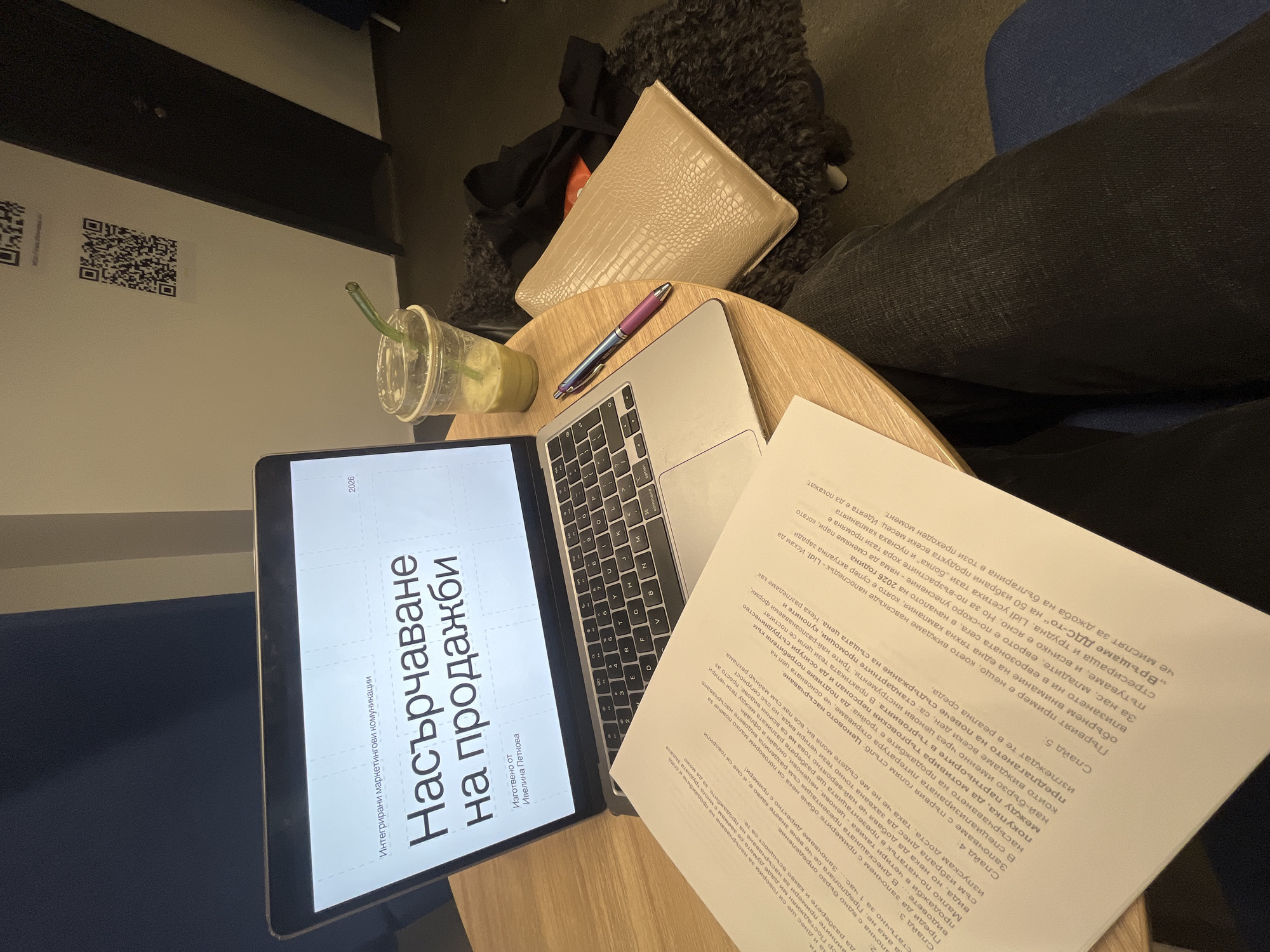 Here is a forensic description of the image:
The image shows a person sitting at a small, round table in what appears to be a café or co-working space. The person has a laptop open in front of them, displaying a presentation slide in Bulgarian titled "Насърчаване на продажби" which translates to "Sales Promotion". Next to the laptop is a stack of printed documents, also in Bulgarian. A pen rests on the laptop near the keyboard, and a plastic cup with a green liquid (possibly a matcha latte) is nearby. A light-colored, textured purse and some dark-colored clothing are placed near the edge of the table, suggesting the person's belongings. The person is wearing dark jeans. In the background, a wall with two QR code stickers is visible.
Based on the presentation title and the text on the printed documents, the subject appears to be related to marketing or business. Due to the text being in Bulgarian, it is highly likely the location is in Bulgaria, specifically Sofia as that is its Capital.