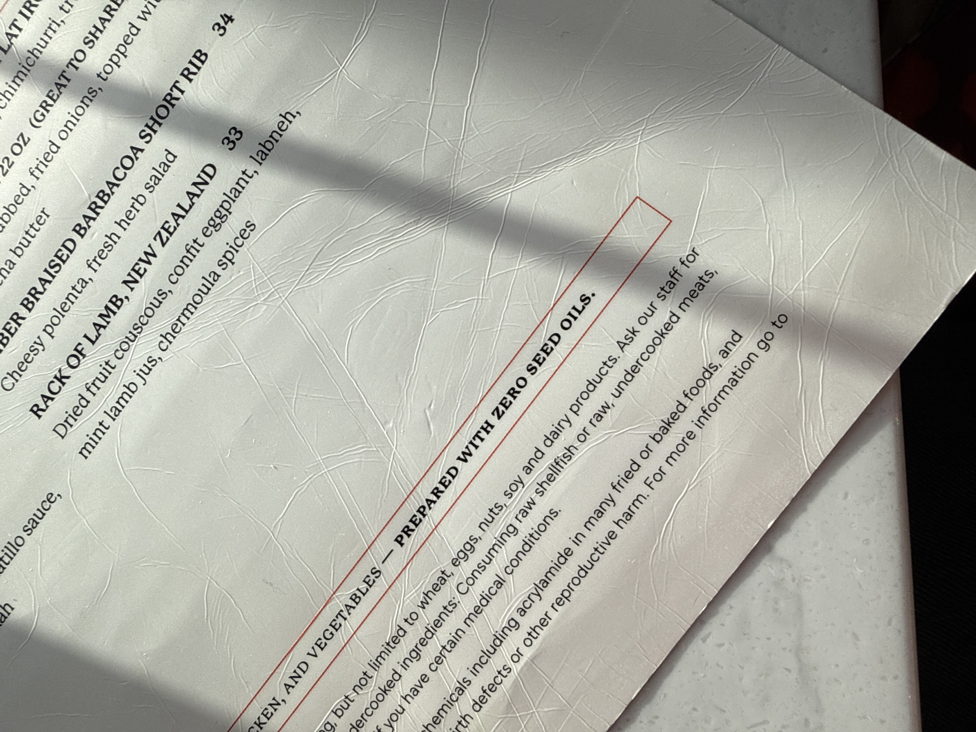 The image displays a close-up, angled view of a white restaurant menu, likely captured indoors. A significant diagonal shadow cuts across the top and middle sections of the menu.
The menu lists several dishes, including "BER BRAISED BARBACOA SHORT RIB" for 34, described with "Cheesy polenta, fresh herb salad." Another item, "RACK OF LAMB, NEW ZEALAND," is priced at 33 and accompanied by "Dried fruit couscous, confit eggplant, labneh, mint lamb jus, chermoula spices." Other partially visible menu entries mention "chimichurri," "fried onions," and "butter."
A key section, highlighted by red lines, declares "CKEN, AND VEGETABLES — PREPARED WITH ZERO SEED OILS." Below this, a comprehensive consumer advisory is present. It states that items may contain allergens "not limited to wheat, eggs, nuts, soy and dairy products" and advises patrons to "Ask our staff" for information, especially if they "have certain medical conditions." The advisory also warns about the risks of "Consuming raw shellfish or raw, undercooked meats." Additionally, a warning about "chemicals including acrylamide in many fried or baked foods" that can cause "birth defects or other reproductive harm" is visible, directing users to a source for more information.
The menu material appears to be a textured, off-white paper or synthetic material, with some minor creases visible across its surface. The overall impression is that of a detailed and informative restaurant menu from a dining establishment in Solana Beach, USA.