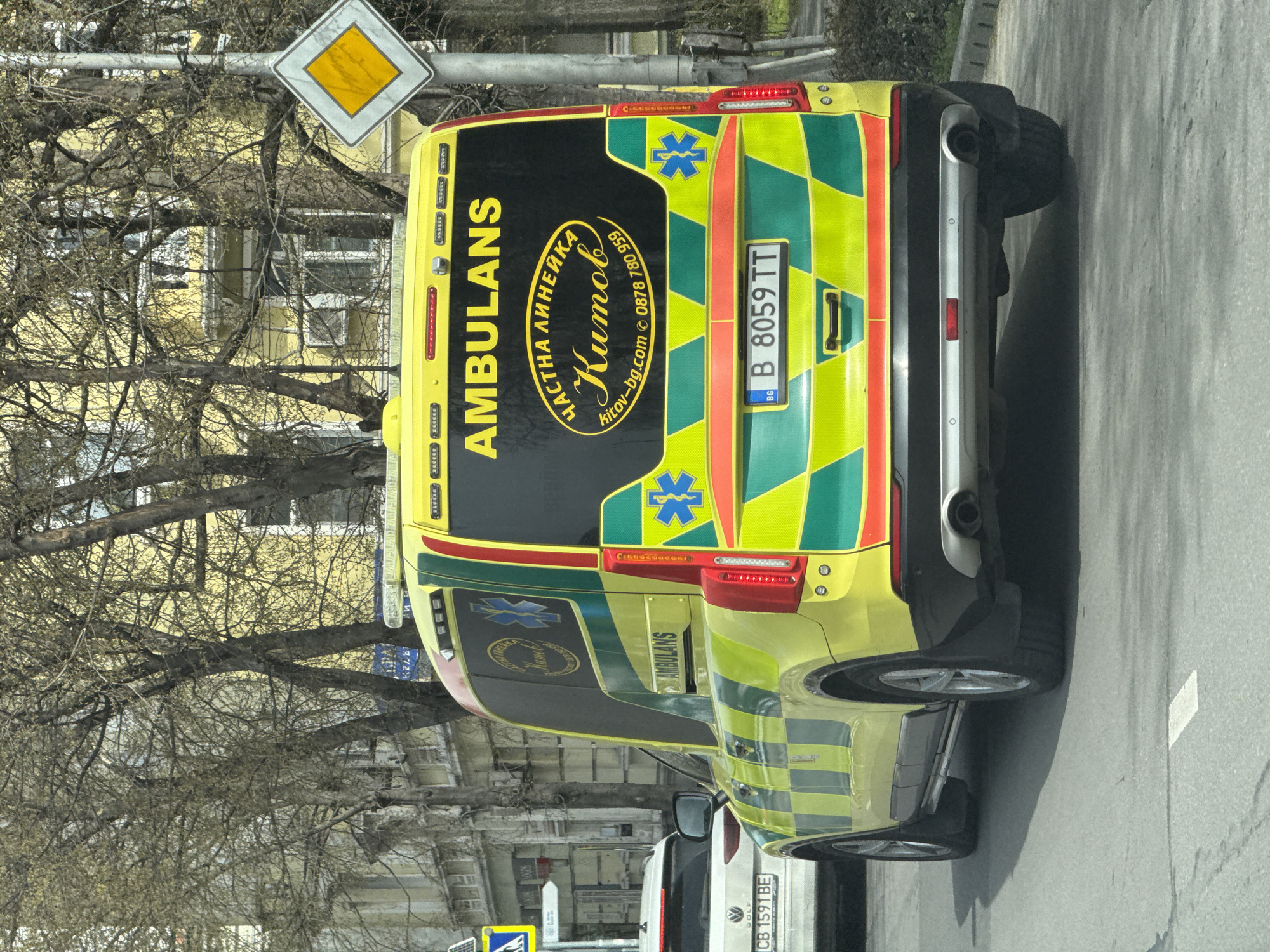 The image shows the rear of two ambulances, likely in Varna, Bulgaria, based on the "B" on the license plate, the Bulgarian web address (kitov-bg.com), and Bulgarian writing on the vehicle. The first ambulance, closer to the camera, has a distinctive yellow and green checkered pattern with the word "AMBULANS" written vertically. The license plate reads "B 8059 TT." There is a sign above that is partially obscured by tree branches. The second ambulance is visible behind the first. A Volkswagen is also visible with license plate CB 1591 BE. The background shows buildings and trees.