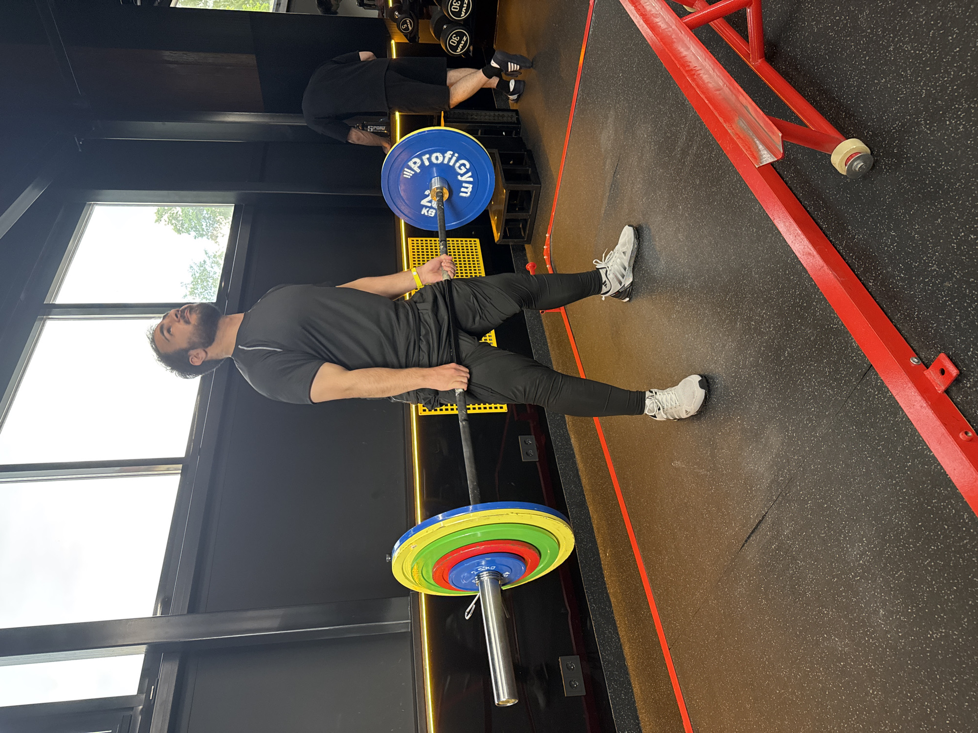 A man with dark hair and a beard, dressed in a black athletic shirt, black compression pants, and white athletic shoes with black accents, is shown holding a barbell in a gym. He wears a yellow band on his left wrist. The barbell is loaded with colorful weight plates; on the side closer to the camera, there are small blue, red, green, and yellow plates, while on the other side, a larger blue plate is visible with "ProfiGym" and "20 KG" printed in white.
The man is standing on a black rubber gym floor, holding the barbell in front of his body, suggesting he is either preparing for or executing a weightlifting exercise like a deadlift. In the background, another person, also in dark athletic wear, is partially visible near a black weight rack containing dumbbells labeled "20 KG" and "30 KG".
The gym features black walls and large windows on the left, letting in natural light and showing an overcast sky with some tree foliage outside, indicating it is daytime. A red metal gym structure runs along the right side of the frame, and yellow strip lighting is present on the black wall behind the main subject.