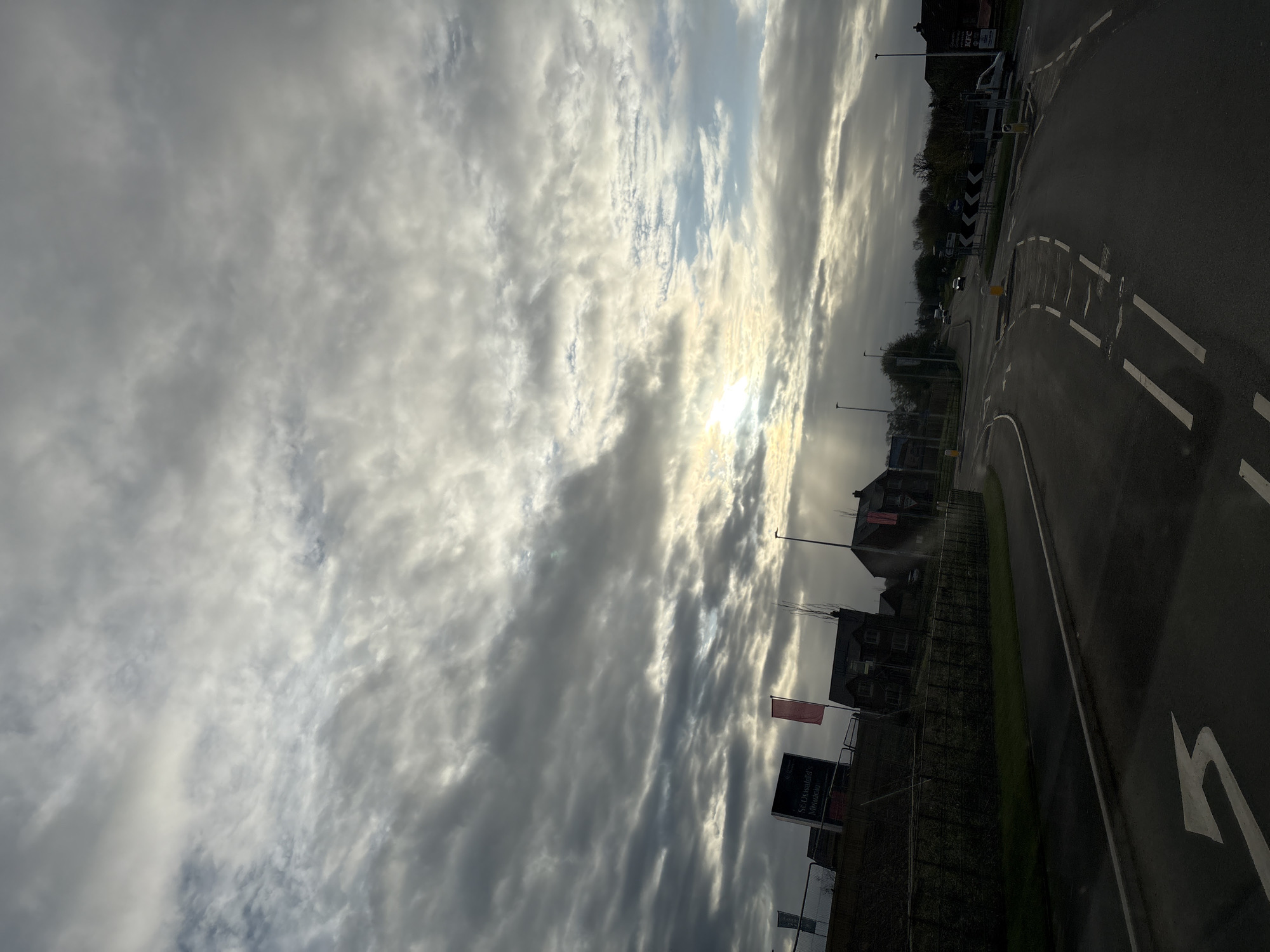 The image captures a view in Oswestry, United Kingdom, featuring a street and cloudy sky. The street, marked with painted road lines and arrows, suggests active traffic flow. Adjacent to the road is a building and perimeter fencing with signage. Overhead, the sky is dominated by thick, layered clouds with some sunlight visible through the gaps.