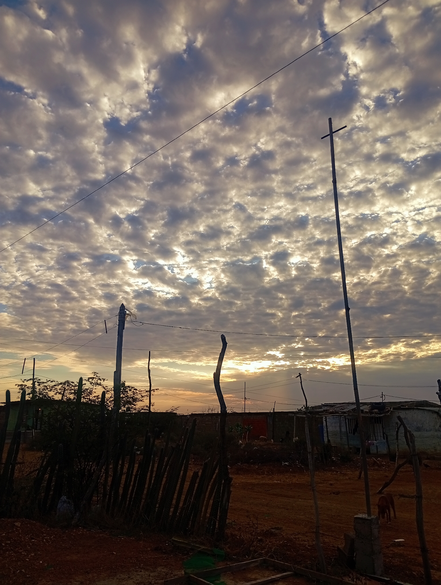 The image depicts a rural scene in Altagracia, Venezuela, at either sunrise or sunset. The sky is filled with altocumulus clouds, partially illuminated by the sun. Power lines run across the upper portion of the frame. A wooden fence made of vertical posts is partially visible in the foreground. Behind the fence are various structures, including simple dwellings constructed of corrugated metal and other materials. A tall, thin metal pole with a cross at the top stands prominently. A dog is visible near one of the structures. The ground is dry and red.